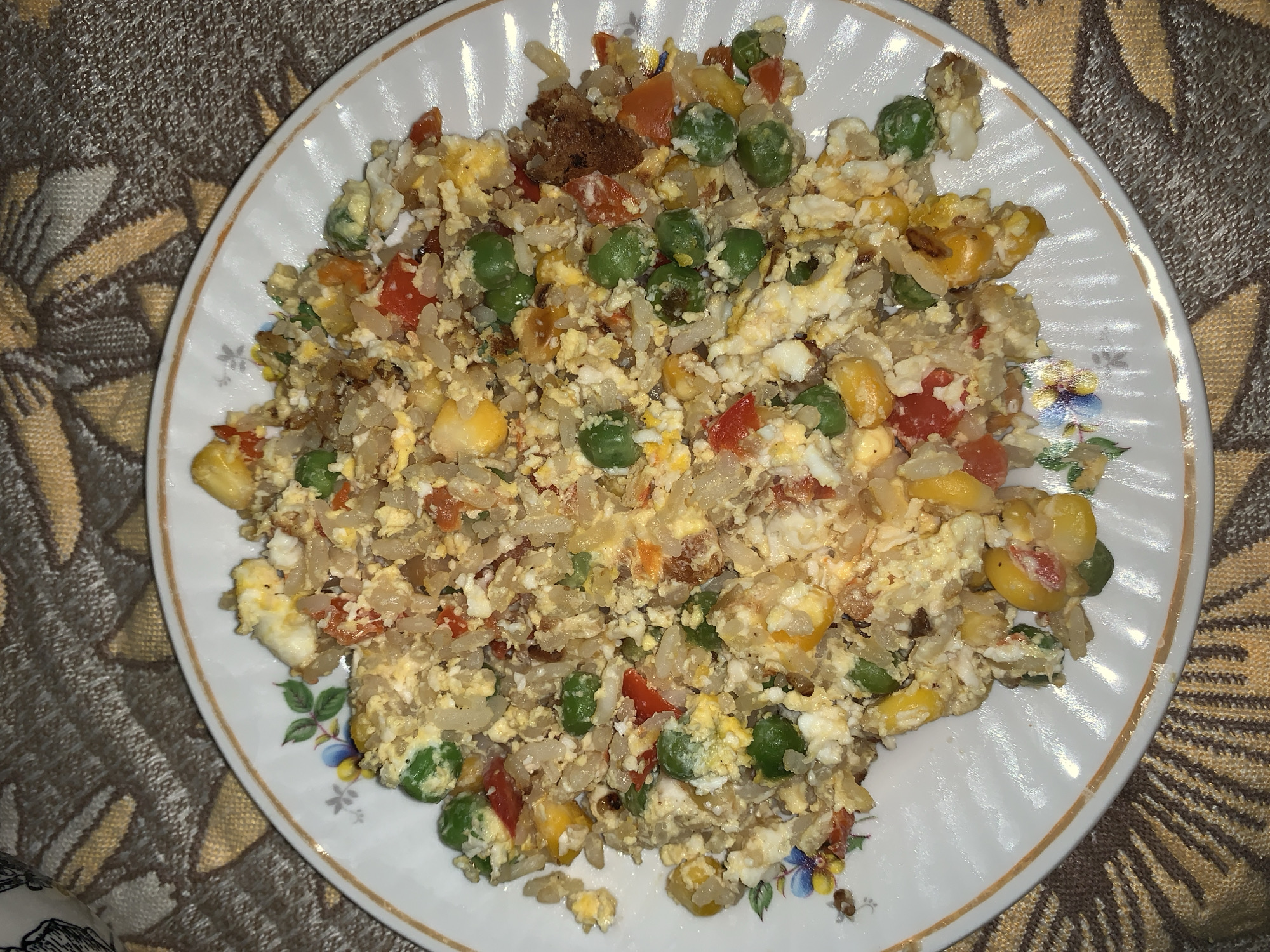 A close-up, top-down shot shows a white, round plate filled with fried rice on a patterned cloth. The fried rice is a mixture of ingredients including scrambled eggs, peas, corn kernels, and diced red bell peppers. The plate has a thin orange rim and a delicate floral pattern along the edge. The cloth beneath the plate features a repeating pattern of brown and tan abstract shapes. The lighting is somewhat dim, with a slight yellow cast.