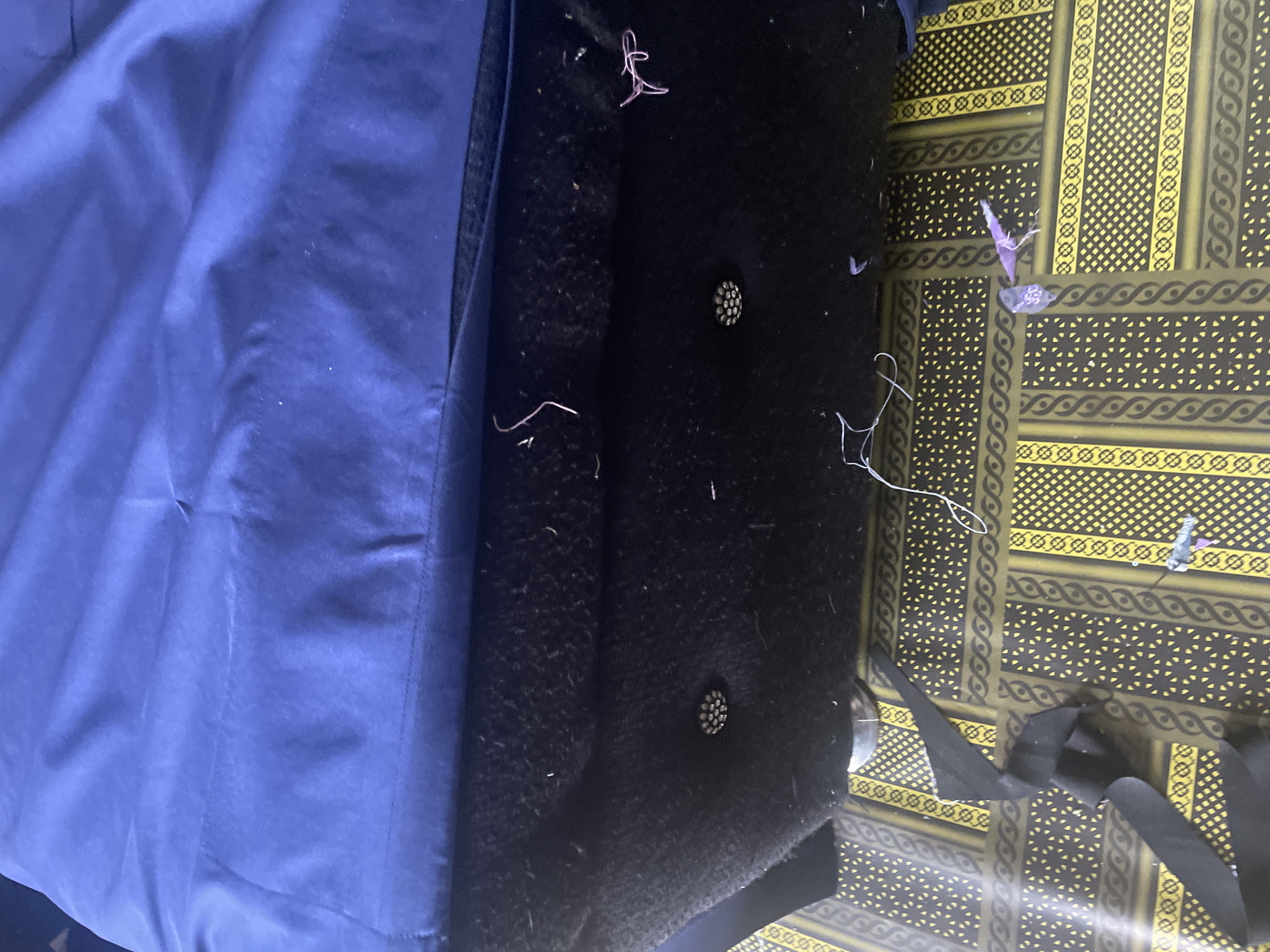 This media file captures a close-up view of a dark fabric, likely a jacket lining, with some loose threads and a few small, purple, plastic-like pieces. The fabric appears to be soft and possibly fuzzy. To the left, a section of a shiny, blue material is visible, possibly the exterior of the same jacket. The blue material has wrinkles and folds, suggesting it is flexible. The scene is set against a patterned background, which is yellow and dark green, with intricate geometric designs.
There are no people visible in the image, nor is any specific activity taking place. The notable details include the loose threads, which might indicate the garment is new or in need of repair, and the small plastic pieces, which could be remnants from packaging or a previous repair. The lighting suggests an indoor setting, and there are no cues about the time of day or weather. The patterned background might offer a clue to the location, but without further context, it's difficult to pinpoint definitively, although the prompt provides a location context of Jalingo, Nigeria. No visible text is present in the media file.