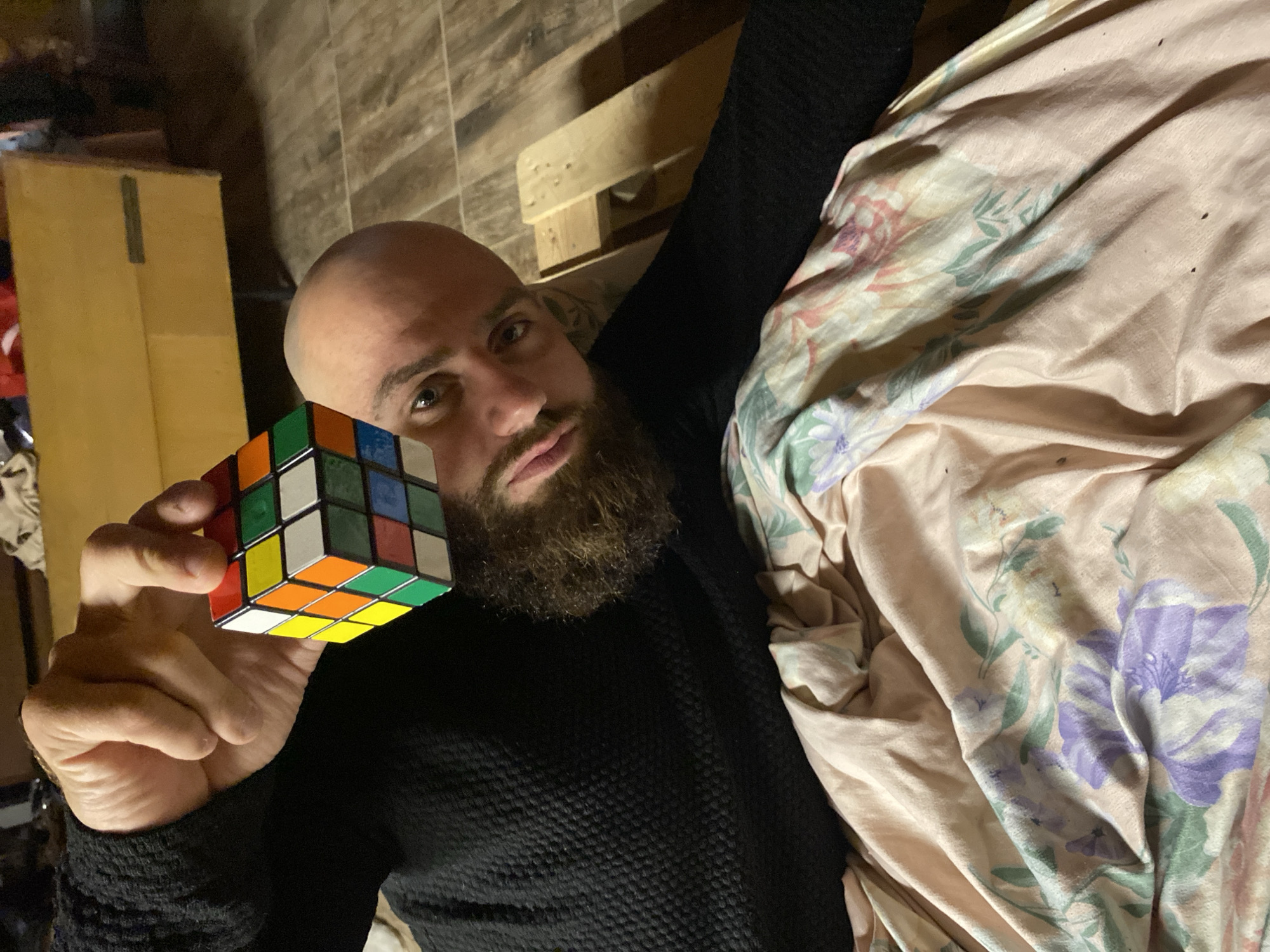 An overhead, slightly angled shot captures a bald, bearded man lying down indoors, looking directly at the camera with a neutral expression. He wears a dark, textured long-sleeved shirt. In his left hand, he holds an unsolved Rubik's Cube, displaying a mix of red, yellow, green, blue, orange, and white squares across its visible faces. To his right, a light peach-colored blanket or sheet with a floral pattern featuring purple, green, and white elements is draped. The background on the left includes a plain cardboard box, while the area behind his head shows wooden planks or panels, possibly part of a wall or floor structure. The lighting is somewhat dim, suggesting an indoor evening or low-light setting. No discernible text is visible in the image. The scene is set in Bankya, Bulgaria.