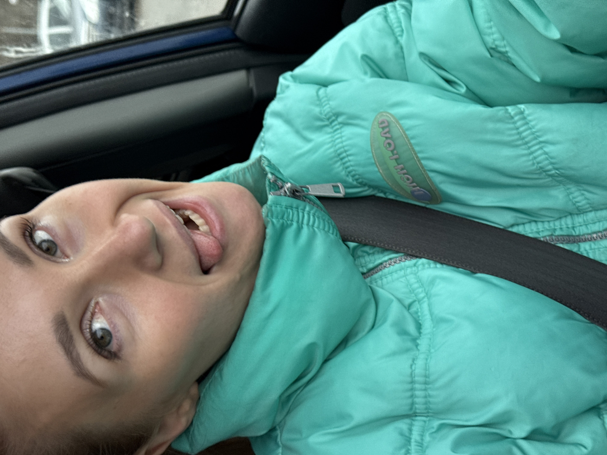 A girl is photographed in a car, wearing a turquoise puffer jacket and a seatbelt. She is sticking her tongue out. The car's interior, including the door frame, is visible. The location is unidentifiable, and there's no indication of Chaikovsky, Russia.