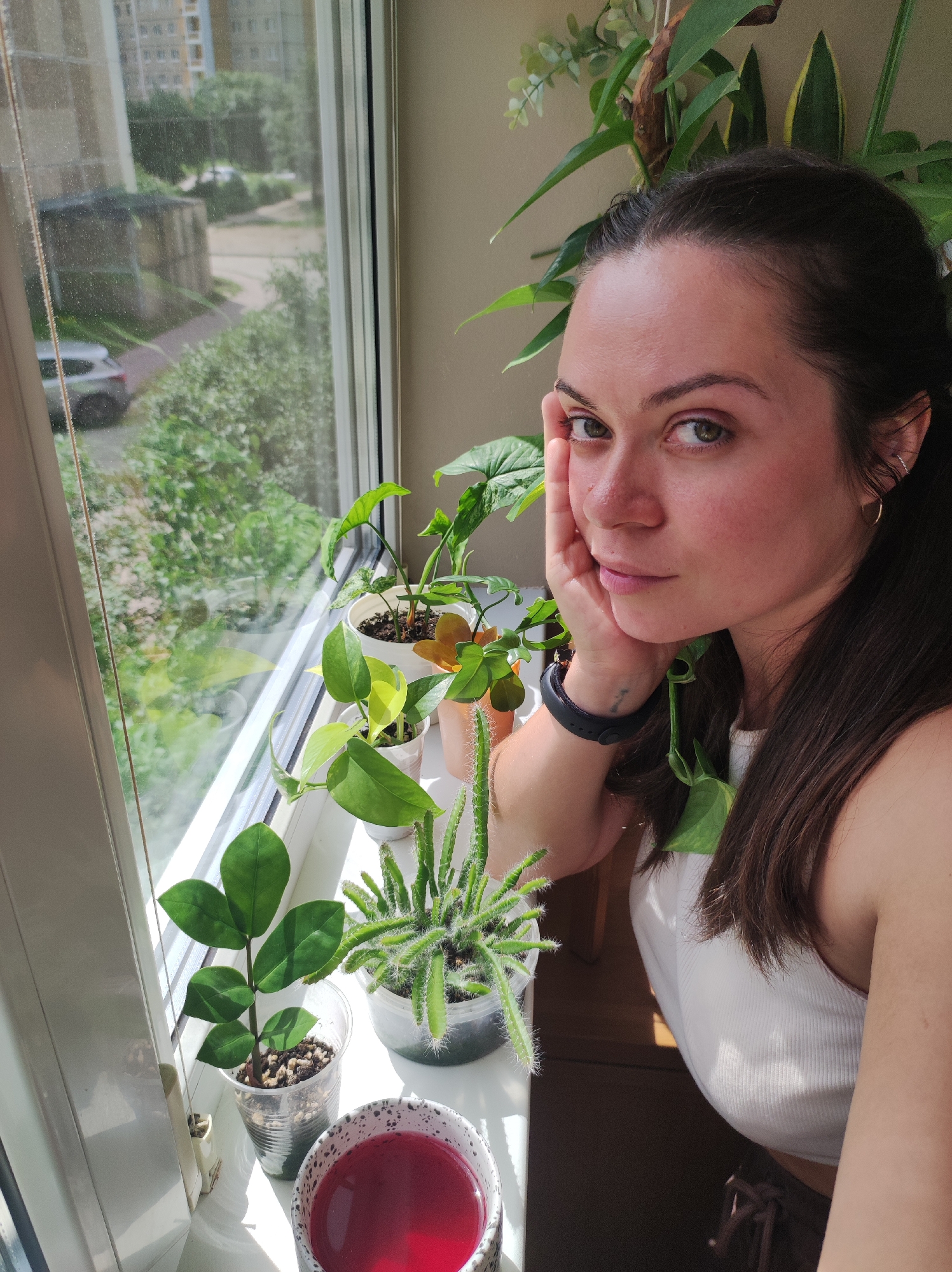 A young woman with dark hair and green eyes is leaning on her hand, looking towards the camera. She is positioned near a window with several potted plants on the windowsill. Outside the window, there is a view of a residential building, trees, and a parked car, suggesting an urban setting. The lighting indicates it is daytime. The woman is wearing a white tank top and a black watch. The scene is indoors, by a window, with a focus on the woman and the plants. The location is identified as Chaikovsky, Russia.