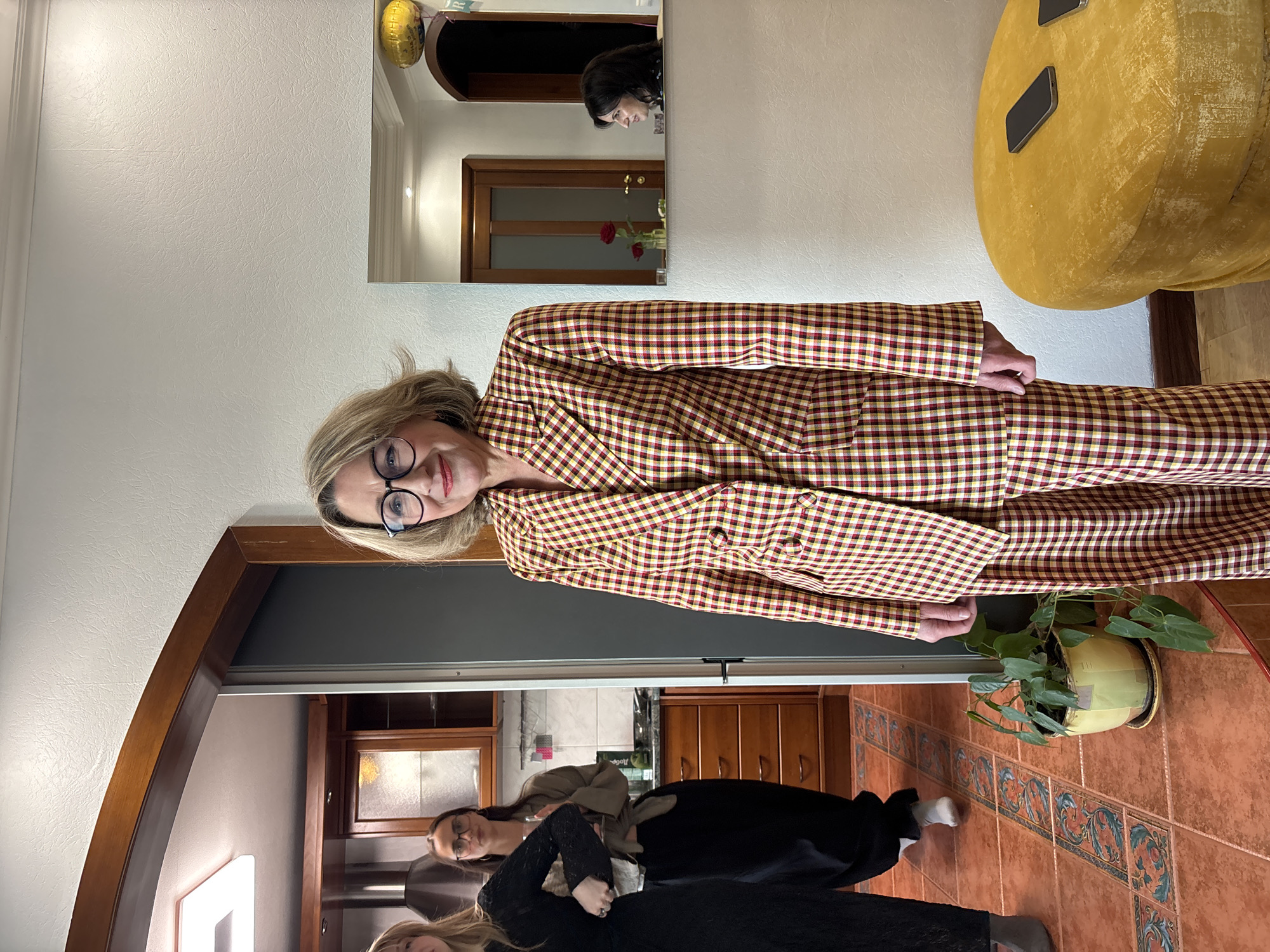 A medium shot depicts an adult woman with blonde hair and eyeglasses, wearing a patterned yellow and red blazer, in a domestic indoor setting. She appears to be standing in a doorway. Behind her, in another room with brown tiled flooring, are two younger women. One is seated, while the other is leaning over her, their faces partially obscured. In the background, there are cabinets and a counter.
In the main room, a large yellow ottoman with a smartphone on top is visible, partially cut off in the upper right corner. A mirror is affixed to the wall, reflecting another woman with dark hair in a different room with a rose on a table. A potted plant stands near the doorway.
There is no indication that the location is in Krasnoyarsk, Russia.
