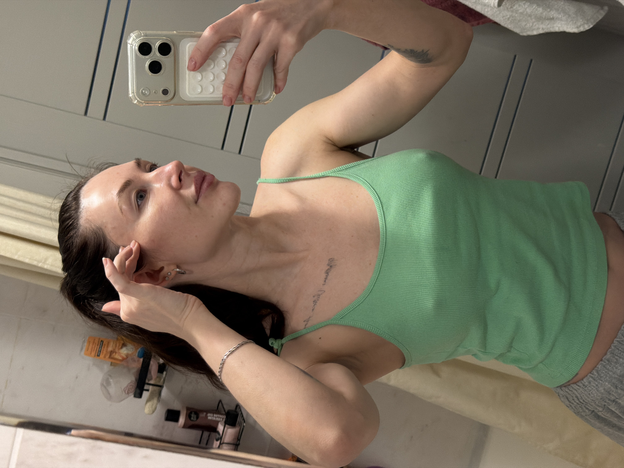 A fair-skinned woman is taking a selfie in a bathroom. She's holding a smartphone with a clear case and a white, textured back. She's wearing a light green ribbed tank top and grey sweatpants. Her dark hair is pulled back, and she's wearing a silver bracelet. Her right hand is touching her face near her ear. There's a shelf with various toiletries and products in the background, suggesting a domestic setting. The background also includes a door with vertical panels and part of a shower curtain. The location is not determinable as Perm, Russia.