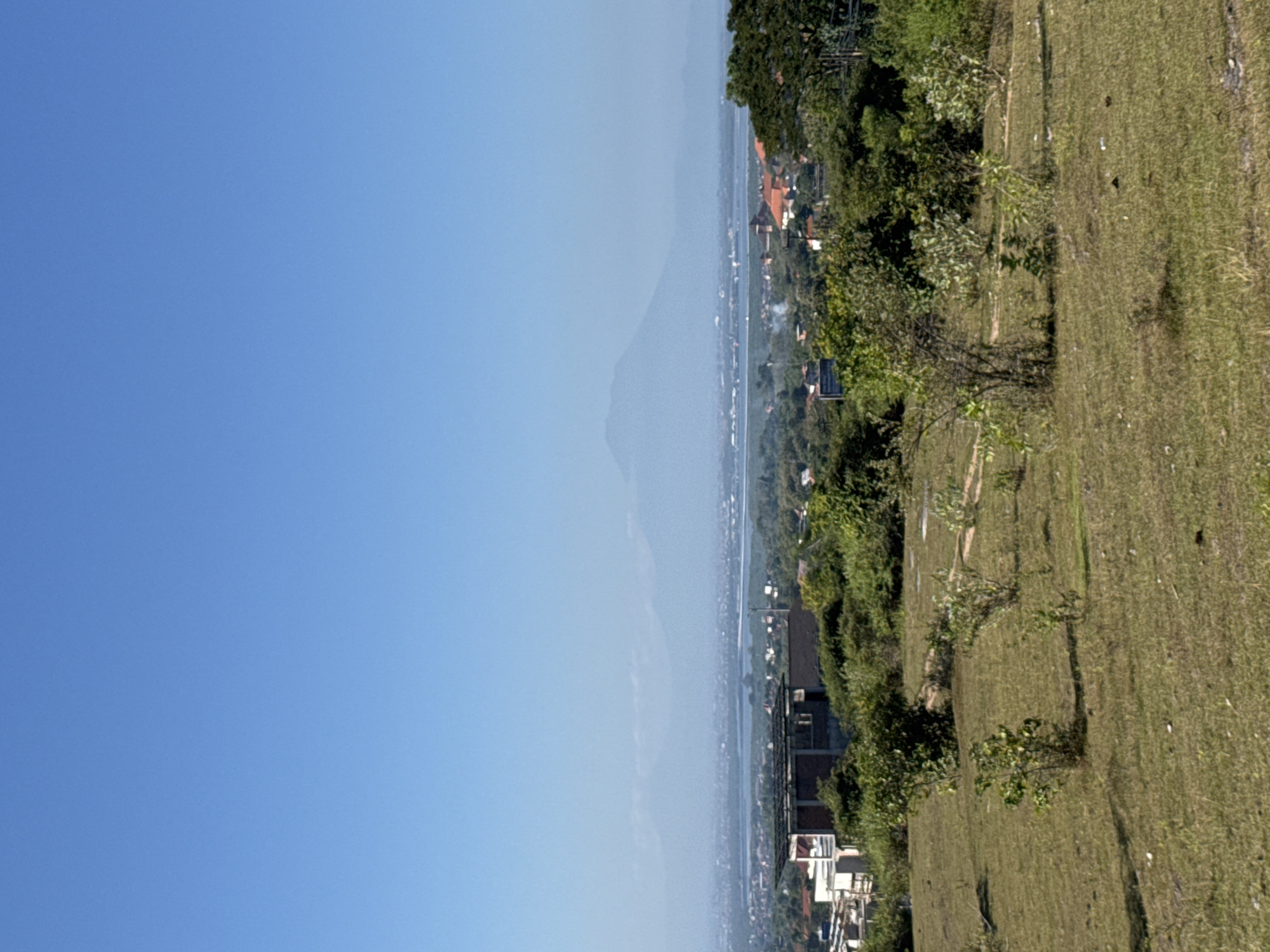 A wide-angle, high-angle shot captures a panoramic view of the Kuta Selatan region in Bali, Indonesia. In the foreground, a grassy, uneven terrain dotted with small shrubs and sparse trees slopes gently downward. The middle ground reveals a densely vegetated area transitioning into a sprawling cityscape. Buildings with terracotta-tiled roofs are interspersed with lush greenery, suggesting residential or commercial development. A body of water, possibly a bay or inlet, separates the cityscape from a distant, hazy mountain range that dominates the background. The sky is a clear, bright blue, indicative of a sunny day. The overall scene depicts a tropical landscape where urban development meets natural scenery, characteristic of the Kuta Selatan area.