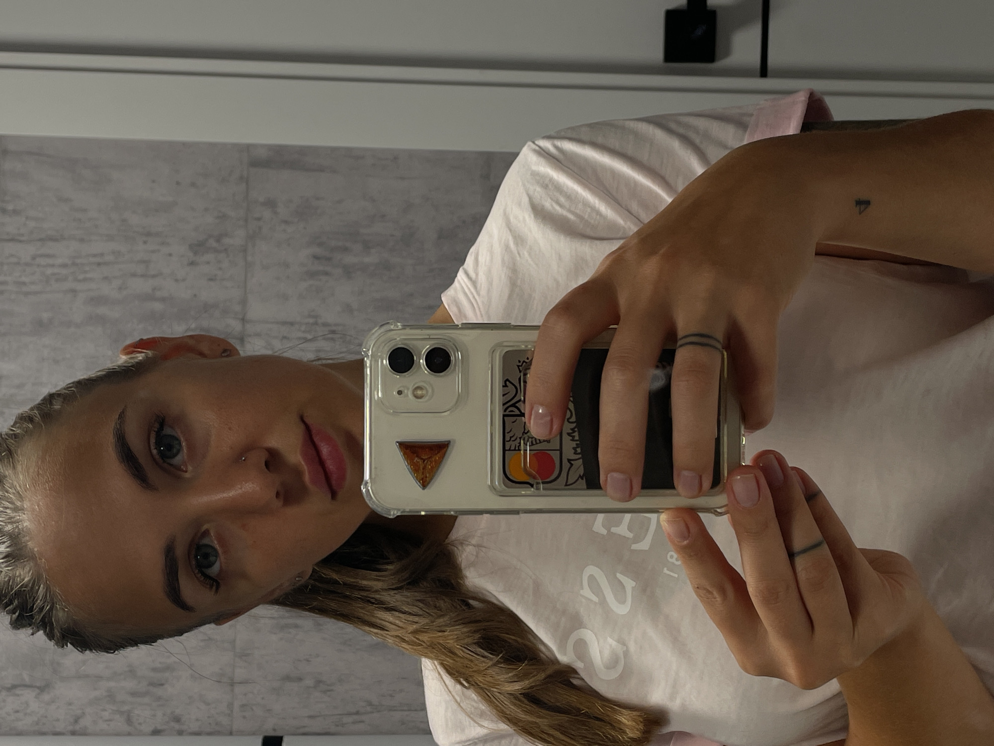 In a modern, tiled bathroom, a young woman with a high ponytail and light blue eyes takes a selfie in a mirror. She is wearing a light pink t-shirt with a white graphic design. She holds a clear phone case with a triangular sticker and a card holder attached. A ring tattoo adorns her left ring finger, and a small triangular tattoo is visible on her right wrist. The lighting is natural, reflecting off the smooth surfaces of the bathroom. No specific landmarks or context suggest the city of Rostov-on-Don.