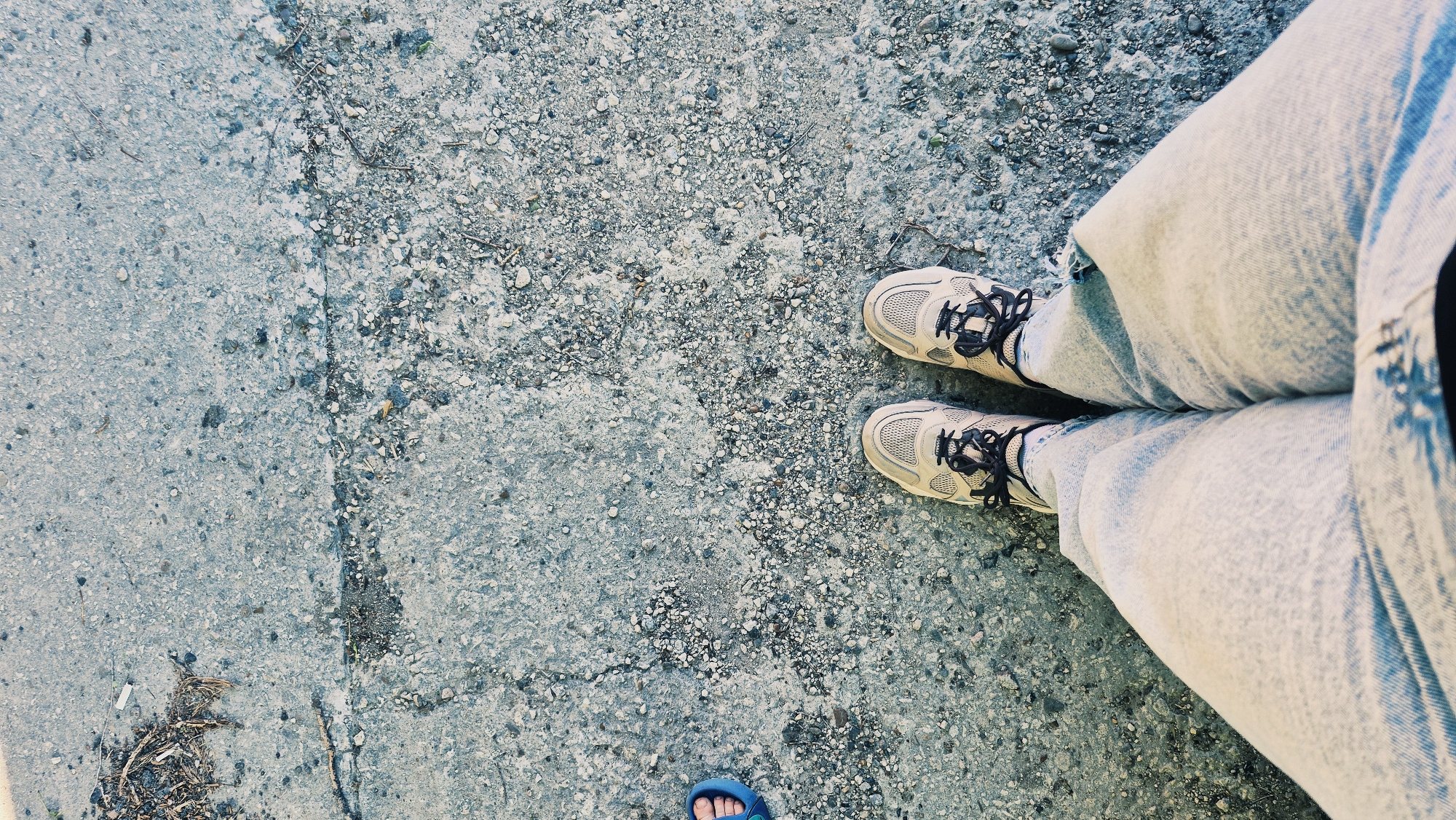 An overhead, full shot captures a person standing on a cracked, textured concrete surface. The person is wearing light-colored jeans and beige athletic shoes with dark laces. To the left of the frame, another person's feet are visible, wearing blue flip-flops. The concrete surface is uneven and contains small pebbles and debris, including some dry leaves in the upper left corner. The lighting suggests it is daytime, with shadows cast on the ground. The scene is devoid of any identifying landmarks that would place it specifically in Chișinău, Moldova.