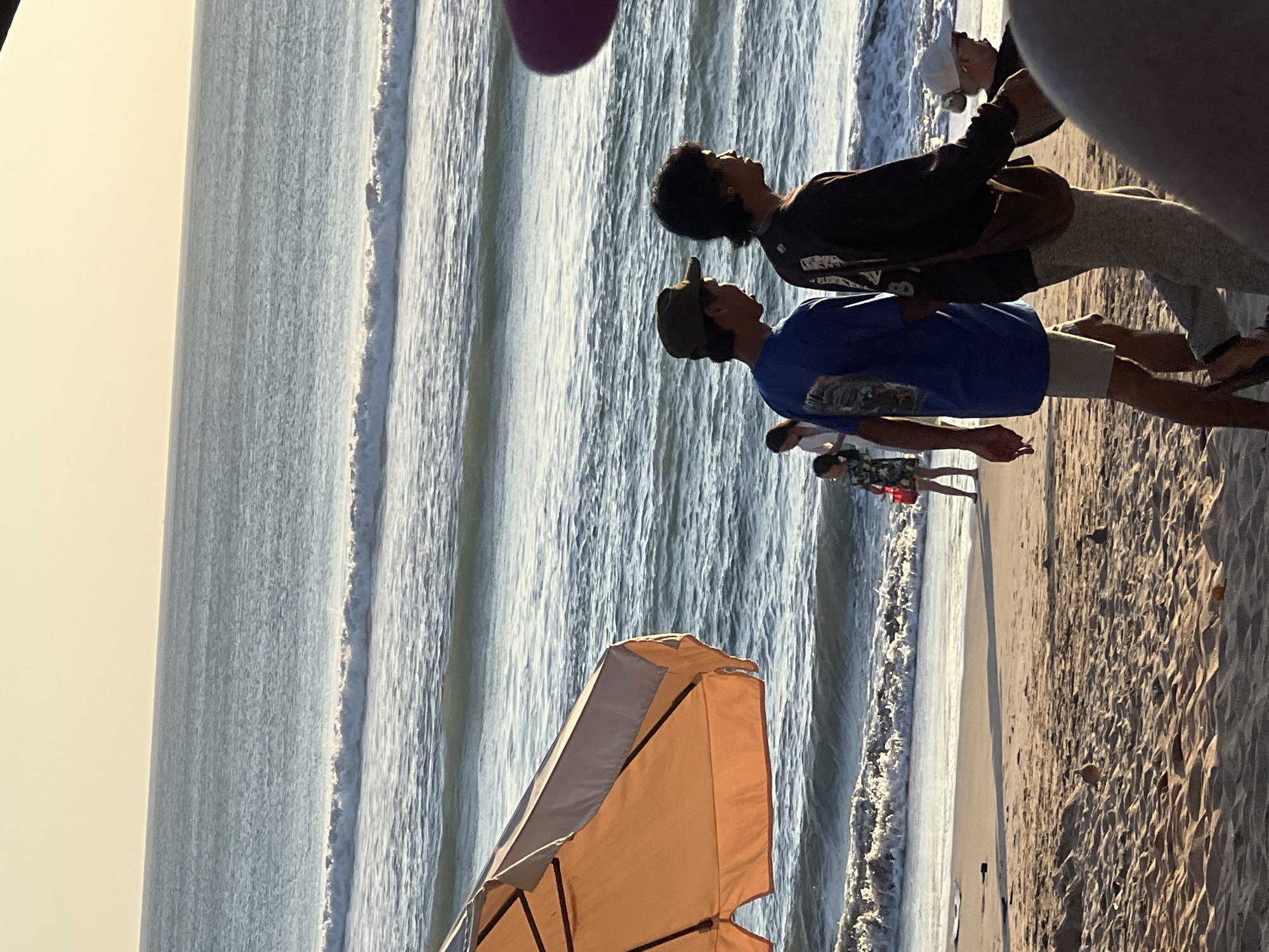 The image depicts a beach scene in Kuta, Indonesia, with the ocean and sky dominating the background. In the foreground, the sandy beach is visible with footprints and scattered debris. An orange beach umbrella is positioned on the left side, partially obscuring the view. Several people are present on the beach. Two young men are walking towards the right, one in a blue t-shirt and khaki shorts, the other in a dark long-sleeved shirt and light-colored pants. Further back, a young girl in a floral dress is walking, holding a red bucket, and a woman in a white dress is also visible. On the far right, another person wearing a white cap is partially visible. The ocean displays gentle waves rolling towards the shore under a clear, pale sky. The lighting suggests it is either early morning or late afternoon.