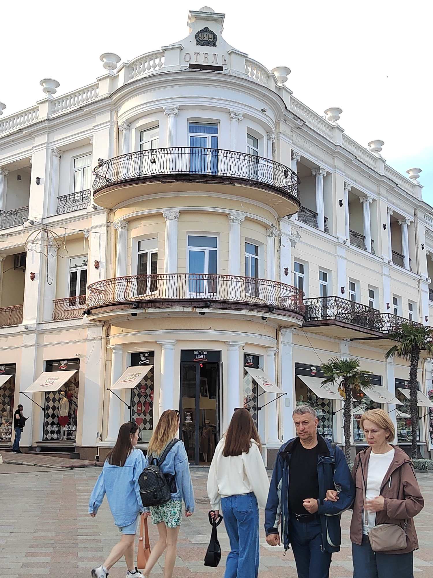 The image captures a street scene in an urban area. A group of four people – three younger women and an older couple – are walking along a paved plaza. The man has his arm around the woman's shoulder.
The background features an ornate, cream-colored building with multiple stories, balconies with wrought-iron railings, and classical architectural elements like columns and decorative urns. The building appears to house commercial establishments, with storefronts displaying awnings and checkered patterns. Signs on the awnings read "DOMINO EIGHT" and appear to advertise couture or fashion-related businesses. Palm trees are planted along the street, adding a touch of greenery. The building's signage reads "ОТЕЛЬ" (Hotel) and "999". A man is standing near one of the storefronts.