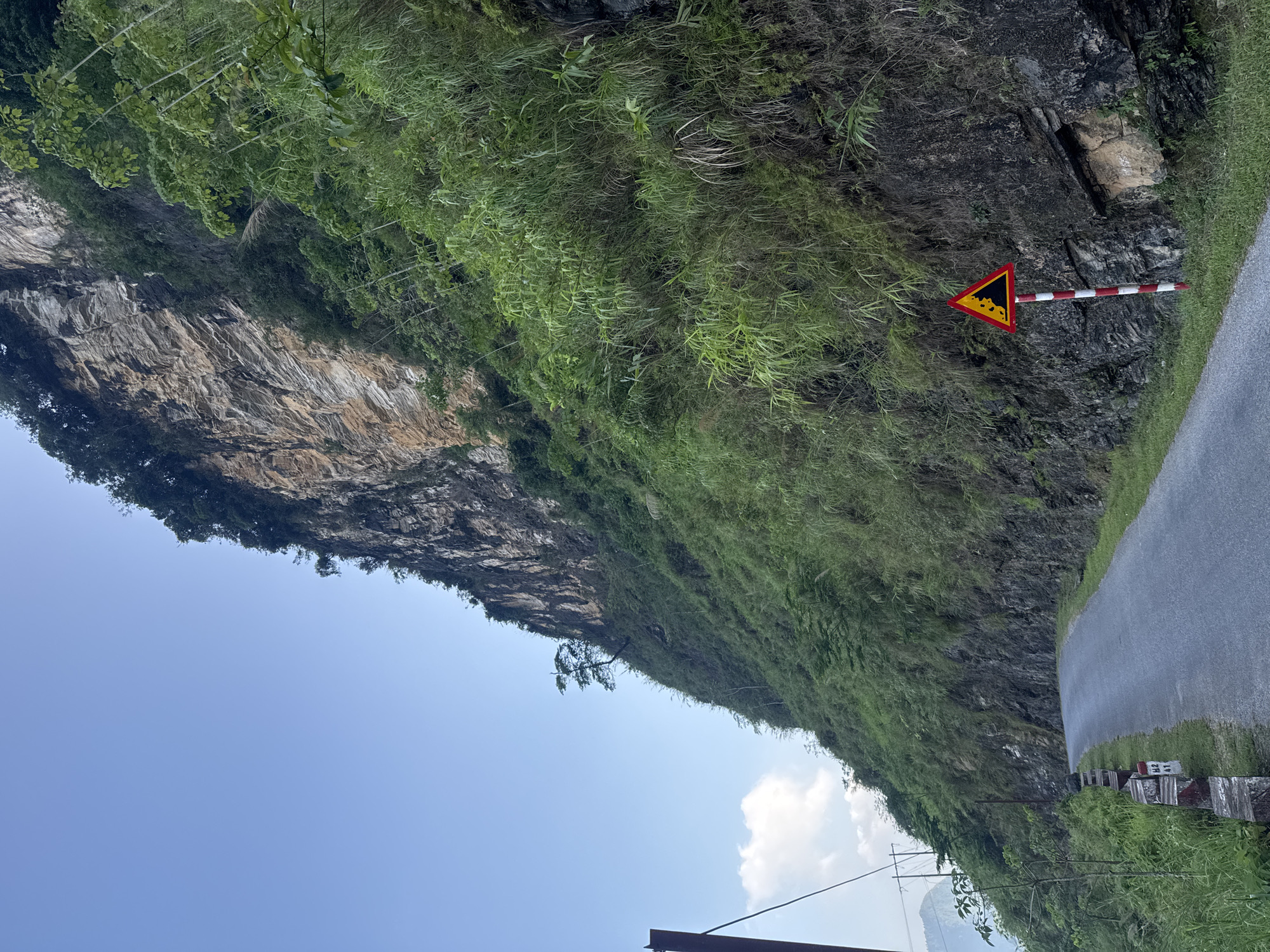 The image depicts a roadside scene in the Đường Thượng Commune, Vietnam. A steep, rocky cliff face covered with lush vegetation towers over a narrow asphalt road. A triangular warning sign with a falling rocks symbol and a red-and-white striped post is positioned on the side of the road. The sky is blue with some scattered clouds.