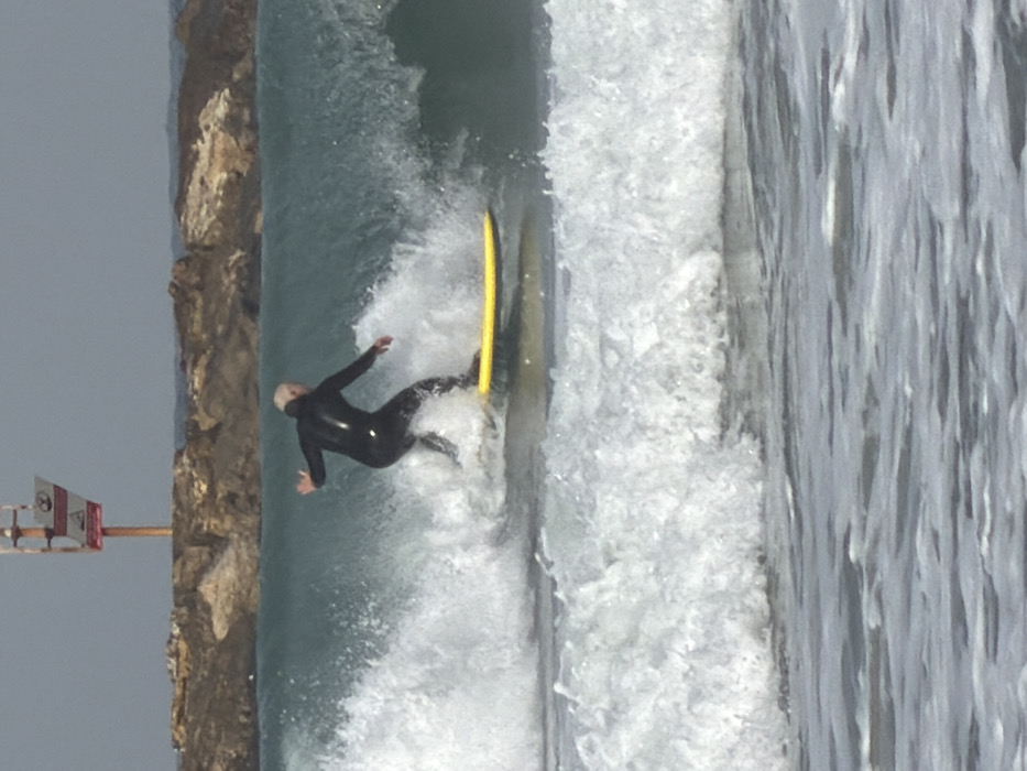The image captures a surfer riding a wave in the Mediterranean Sea off Tel-Aviv, Israel. One individual, an adult, is visible from behind, positioned on a yellow longboard-style surfboard. The surfer wears a black full-body wetsuit and has light-colored hair. The individual is in a right-foot-forward stance, balancing on a prominent breaking wave with white foam and a turquoise face. In the background, a man-made breakwater composed of large rocks spans the horizon. Above and slightly behind the breakwater, a tall pole is erected, featuring at least two rectangular signs: a reddish-orange sign atop a lighter one, consistent with a maritime warning or navigation marker. The sky is overcast and gray. Smaller, white-capped waves are present in the foreground water.