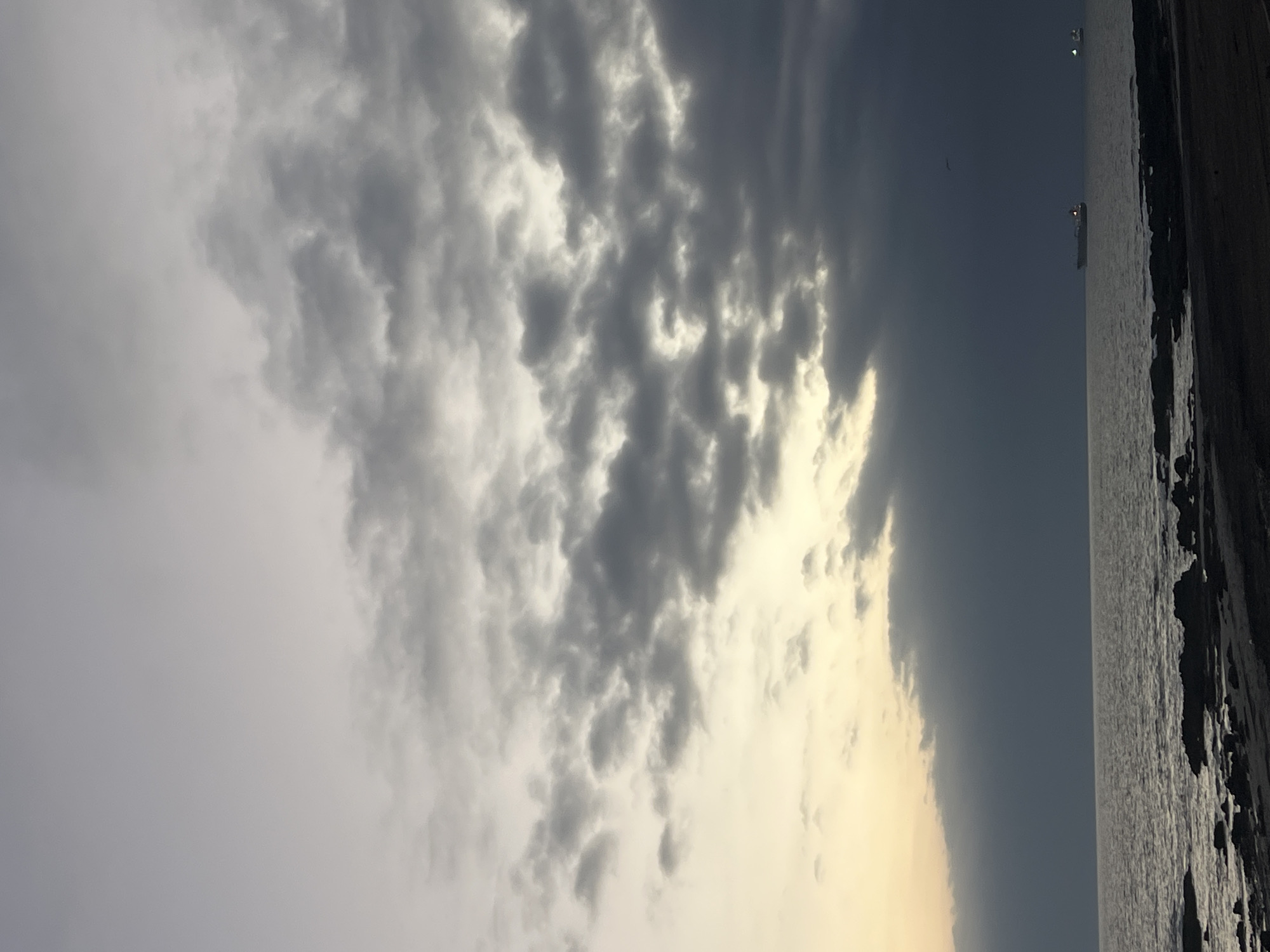 The image shows a view of the coast in Arica, Chile, likely taken at sunset or sunrise. The sky is predominantly covered with clouds, ranging from light grey to darker, more textured formations. The sun is either setting or rising, casting a yellow-orange glow near the horizon where the sky meets the sea. The water surface is relatively calm with two ships on the horizon. The coastline is rocky, with dark, uneven surfaces. The overall scene is serene, capturing the natural beauty of the coastal landscape under dramatic lighting conditions.
