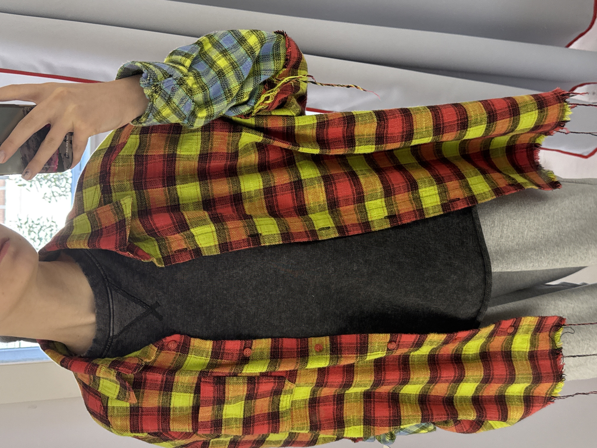 A person is wearing a gray shirt and a yellow and red plaid flannel shirt. The flannel shirt has frayed edges. The person is holding a smartphone in their left hand. The background is blurred, but a window and a white wall are visible. The exact location is Unknown, Unknown.