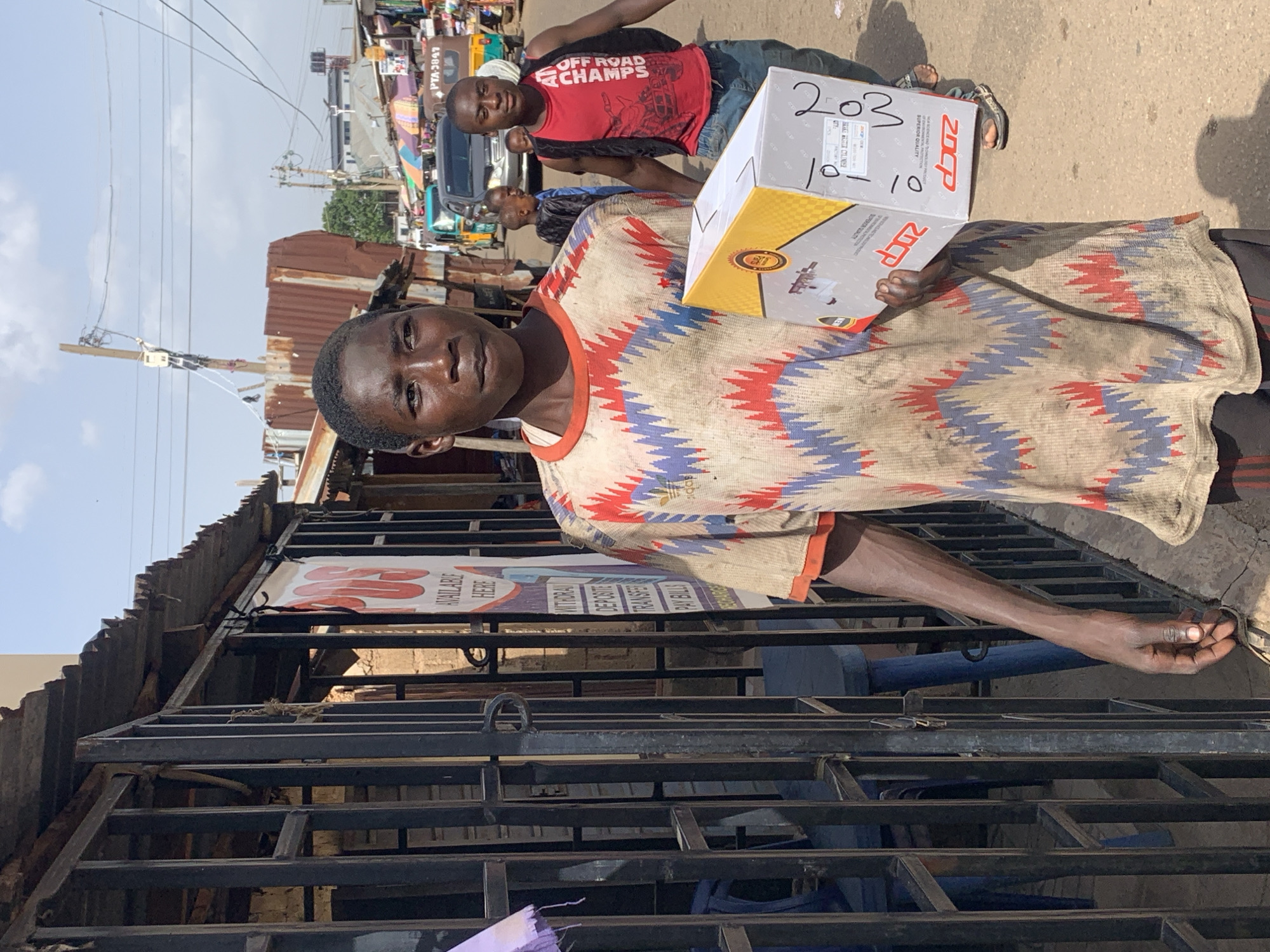 The image depicts a street scene in Jos, Nigeria. In the foreground, a young man of African descent, with dark skin and short black hair, is walking towards the viewer's right, carrying a rectangular cardboard box with yellow and white branding. He is wearing a t-shirt with a red, blue, and beige zigzag pattern, and dark-colored pants. His left arm is bent, holding the box at waist level, and his right arm is partially visible.
Behind him, the environment appears to be a busy marketplace or street. To the left, a dark metal gate with vertical bars encloses a space with blue chairs visible behind it. A sign with red text is partially obscured by the gate. The background shows buildings with corrugated metal roofs, power lines, and other individuals walking. Another young man of African descent, wearing a red t-shirt and a black vest, is walking behind the main subject, also towards the right. A yellow and green motorized rickshaw, commonly known as an "okada," is visible further in the background, suggesting transportation activity. The ground is paved and dusty. The overall lighting suggests a daytime scene under a bright sky.