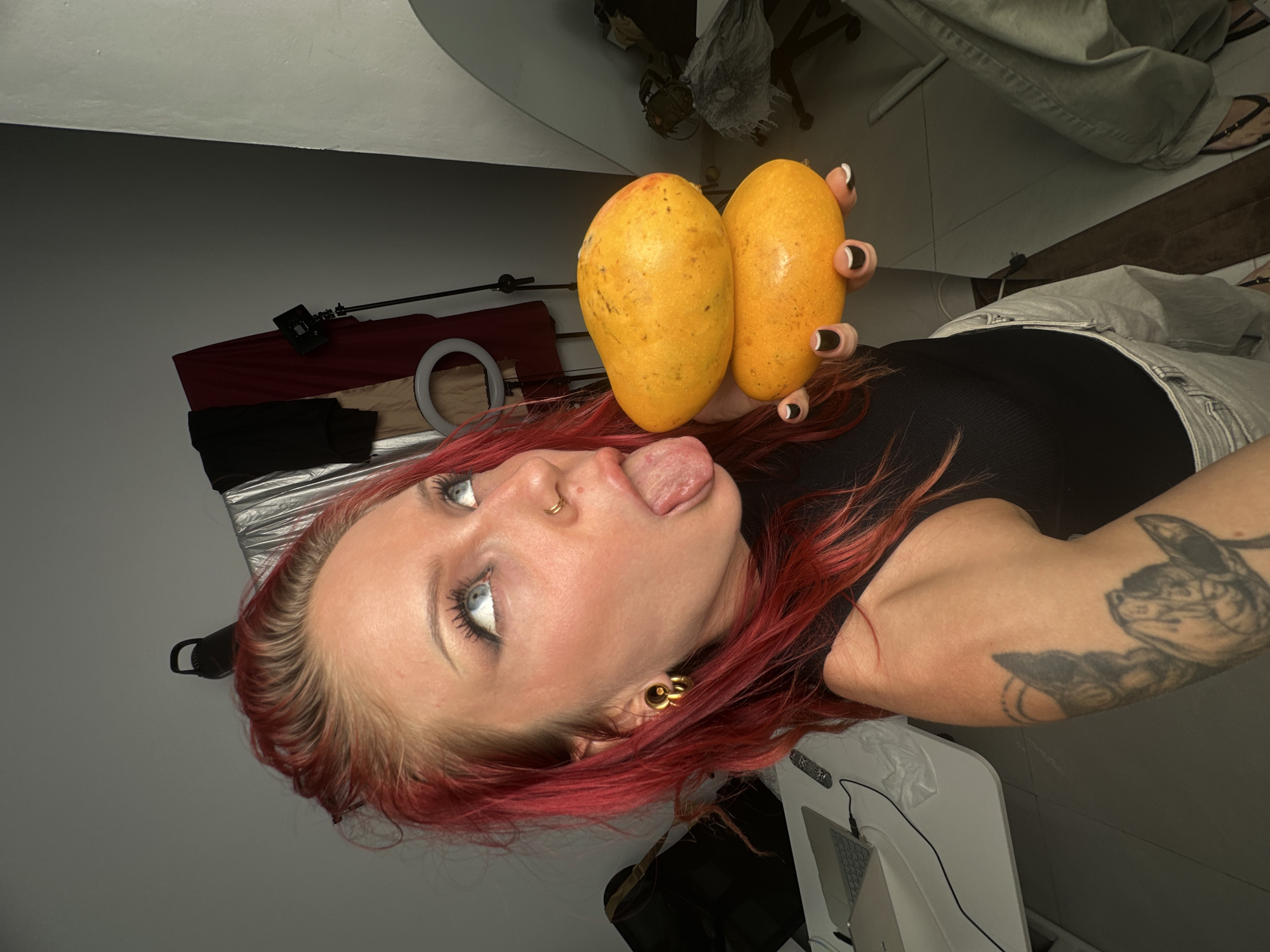 In a dimly lit indoor environment, possibly a studio or a bedroom in North Kuta, Indonesia, a woman with vibrant red hair and blue eyes is the central focus. She is holding two ripe papayas stacked on top of each other in her right hand, her fingers adorned with black and white nail polish. Her tongue is extended, and her eyes are rolled back, giving her a playful or suggestive expression. She is wearing a black tank top and light-colored pants. To her left, a white desk with a laptop and various cables is visible, suggesting a workspace. In the background, draped fabrics and a ring light indicate a setup for photography or videography. The overall impression is one of a candid, personal moment captured in a semi-organized, possibly creative, space.