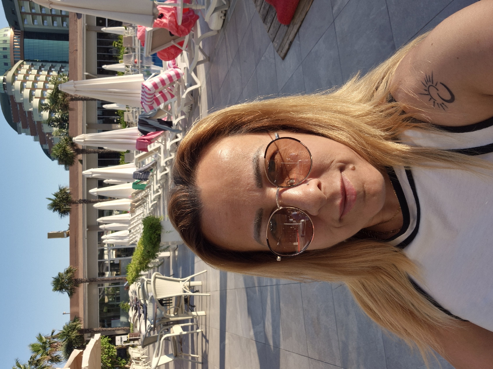 A close-up, selfie-style, medium shot captures a blonde woman wearing sunglasses and a white top with a black trim. She is positioned in the foreground, facing the camera, with her right shoulder and arm visible. A tattoo of a crescent moon and sun symbol adorns her right shoulder. The background reveals an outdoor setting with a paved area, several white lounge chairs, closed white umbrellas, and palm trees. In the distance, a multi-story building with a curved facade and green-tinted windows is visible against a clear blue sky. The lighting suggests it is daytime, likely late afternoon due to the long shadows.