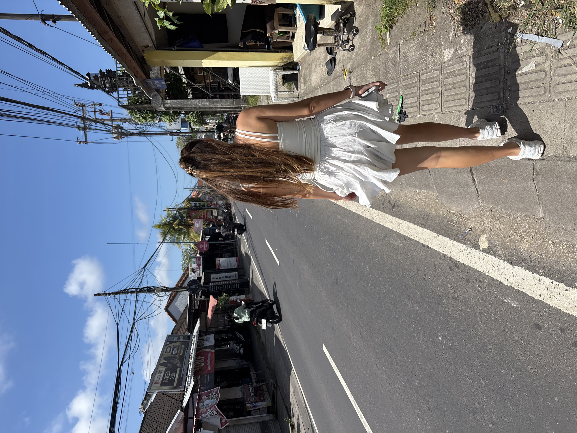 A woman in a white dress and sneakers stands on a sidewalk next to a street in Kuta Selatan, Indonesia. Her back is to the camera. She has long brown hair and is holding a phone in her right hand. The street has white lines down the middle and motorbikes parked on the side. Buildings line the street, displaying advertisements. Power lines run above the street. The sky is blue with scattered clouds.