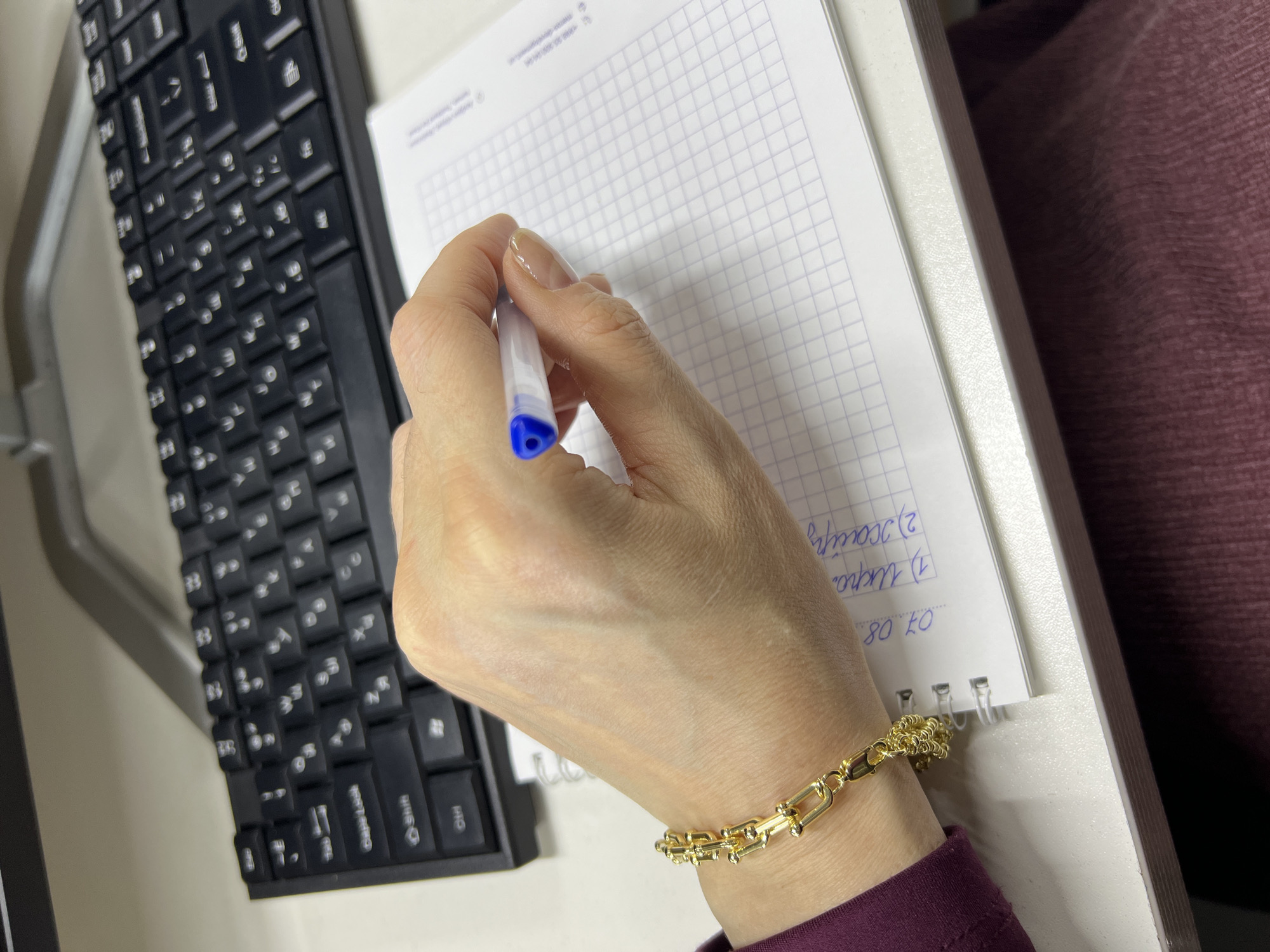 The image shows a close-up view of a person's hand writing on a notebook. The hand, adorned with a gold bracelet, is holding a blue and white pen. The notebook is open to a page with grid lines and some handwritten text in Russian. In the background, a black keyboard is visible, suggesting a desk or office environment. The overall lighting is soft, indicating an indoor setting. There is no visible indication of the city of Shahrixon, Uzbekistan in this image.