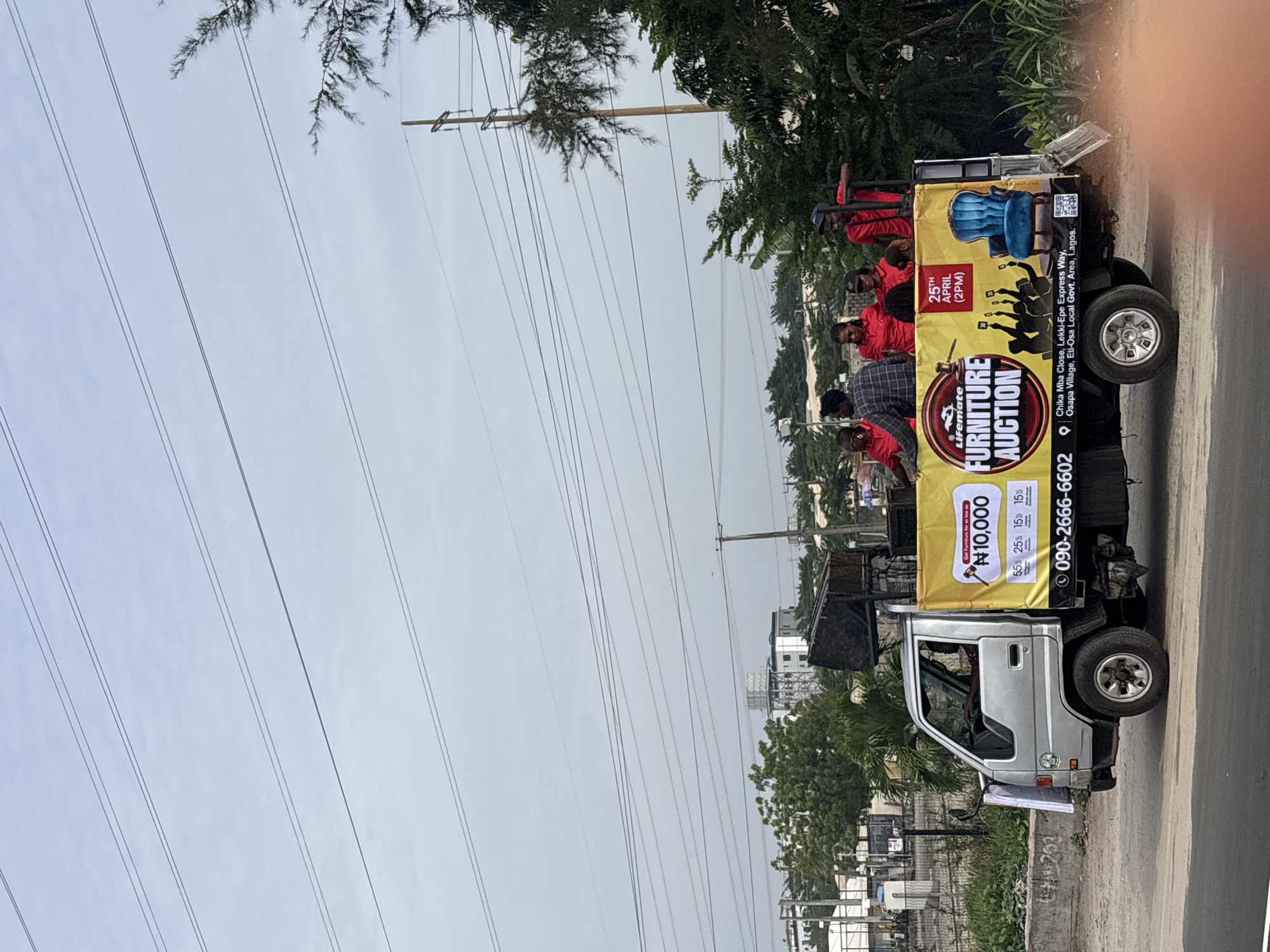 The image captures a silver truck parked on the side of a road in Igboefon, Nigeria, advertising a Lifemate furniture auction. The truck has a large yellow banner on its bed that details the auction, including the date (April 25th, 2 PM) and starting price (₦10,000). The banner also features furniture images and a phone number for contact. Several people are standing on the back of the truck behind the banner. Overhead power lines are visible against a partially cloudy sky. Buildings and trees line the street.