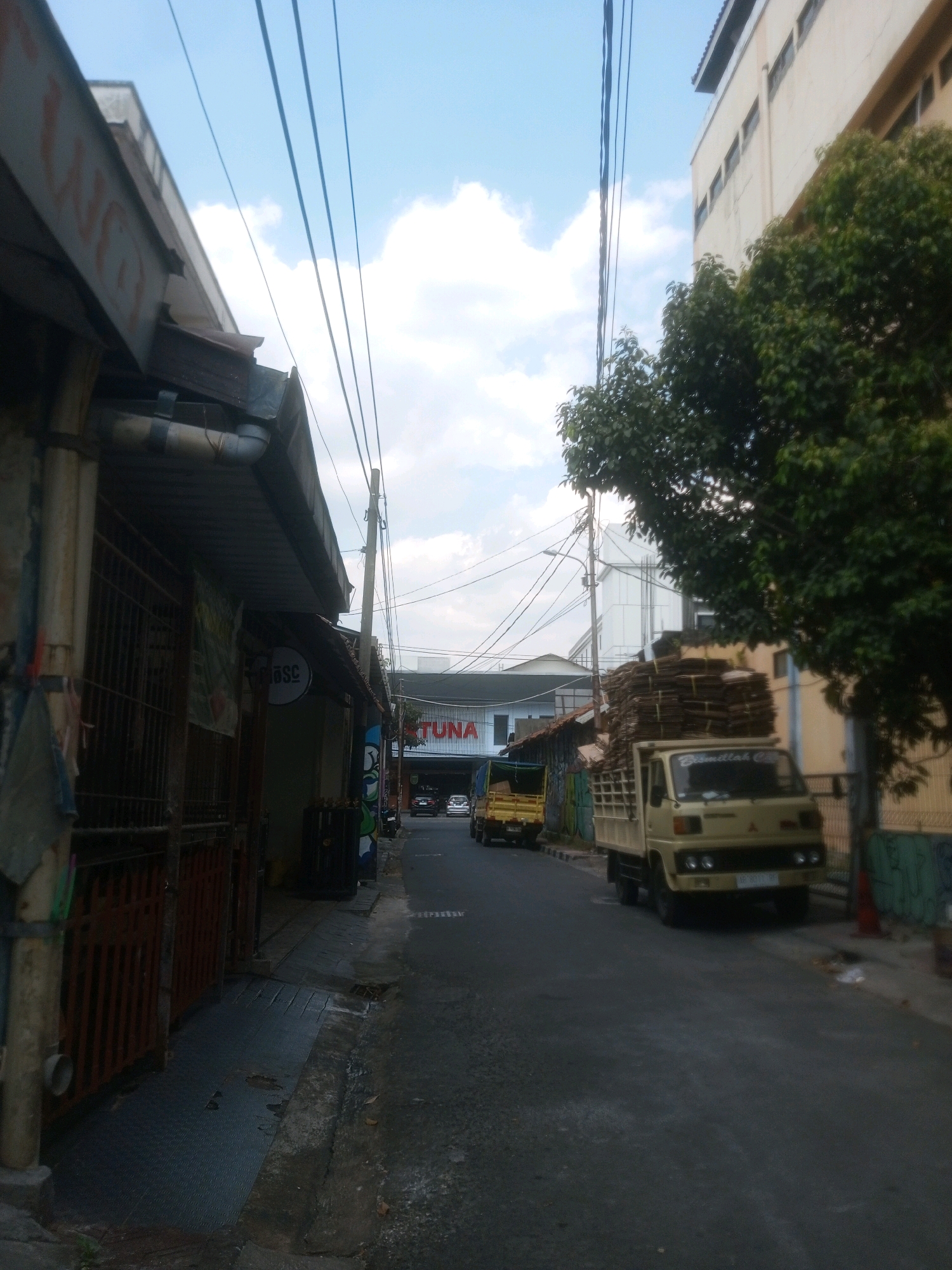 The image captures a street scene in Yogyakarta, Indonesia, under a partly cloudy sky. In the foreground on the left, a row of buildings with awnings and metal gates lines the street. One building has a sign with "BSC" written on it. Further down the street on the right, a beige truck with a load of what appears to be stacked cardboard boxes is parked. Behind it, another yellow truck is visible. A building with the word "TUNA" in red letters is in the background. The street itself is paved with asphalt and shows signs of wear. Electrical wires are strung across the sky, supported by utility poles. Lush green trees are present along the right side of the street, obscuring parts of the buildings. The overall impression is a busy urban environment, likely a commercial or industrial area.