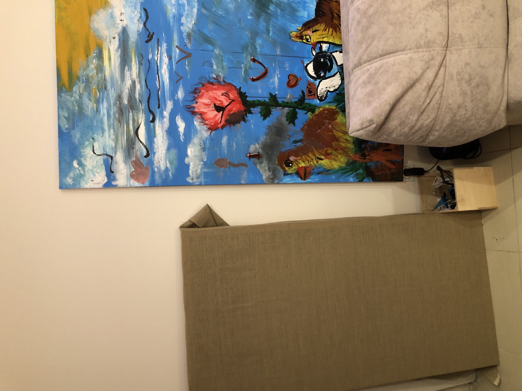 The image depicts an interior scene, possibly a room in a residential apartment or a studio space. A large, unframed canvas leans against a cream-colored wall on the left side of the image. This canvas appears to be made of a coarse, beige fabric, possibly burlap or linen, and is folded at the top right corner.
To the right of the fabric-covered canvas, a colorful, abstract painting hangs on the wall. This painting features a bright blue sky with white and grey clouds, a large red flower with a black center, and several abstract figures that resemble animals, including what appears to be a duckling, a calf, and another bird or chick. There are also black, wavy lines scattered across the sky, possibly representing birds or abstract designs, and a yellow area in the upper right corner suggesting sunlight.
In the foreground, a portion of a light-colored, quilted sofa or ottoman is visible on the right side of the image, partially obscuring the bottom of the painting. In the lower middle section of the image, between the fabric canvas and the painting, a small wooden crate sits on the floor. This crate contains various items, including what appear to be art supplies or tools, and a black electrical cord. A wall socket is visible on the wall behind the crate, with a black electrical plug inserted into it.
The floor appears to be tiled with square, light-colored tiles. The lighting suggests an indoor environment, likely illuminated by artificial light sources, possibly supplemented by natural light coming from an unseen window.
There are no people visible in the image. The scene is static, with no apparent action or interaction occurring. The overall impression is one of an unfinished art space or a room undergoing redecoration. The image does not provide any specific visual cues to indicate its location within the city of Saint Petersburg, Russia.