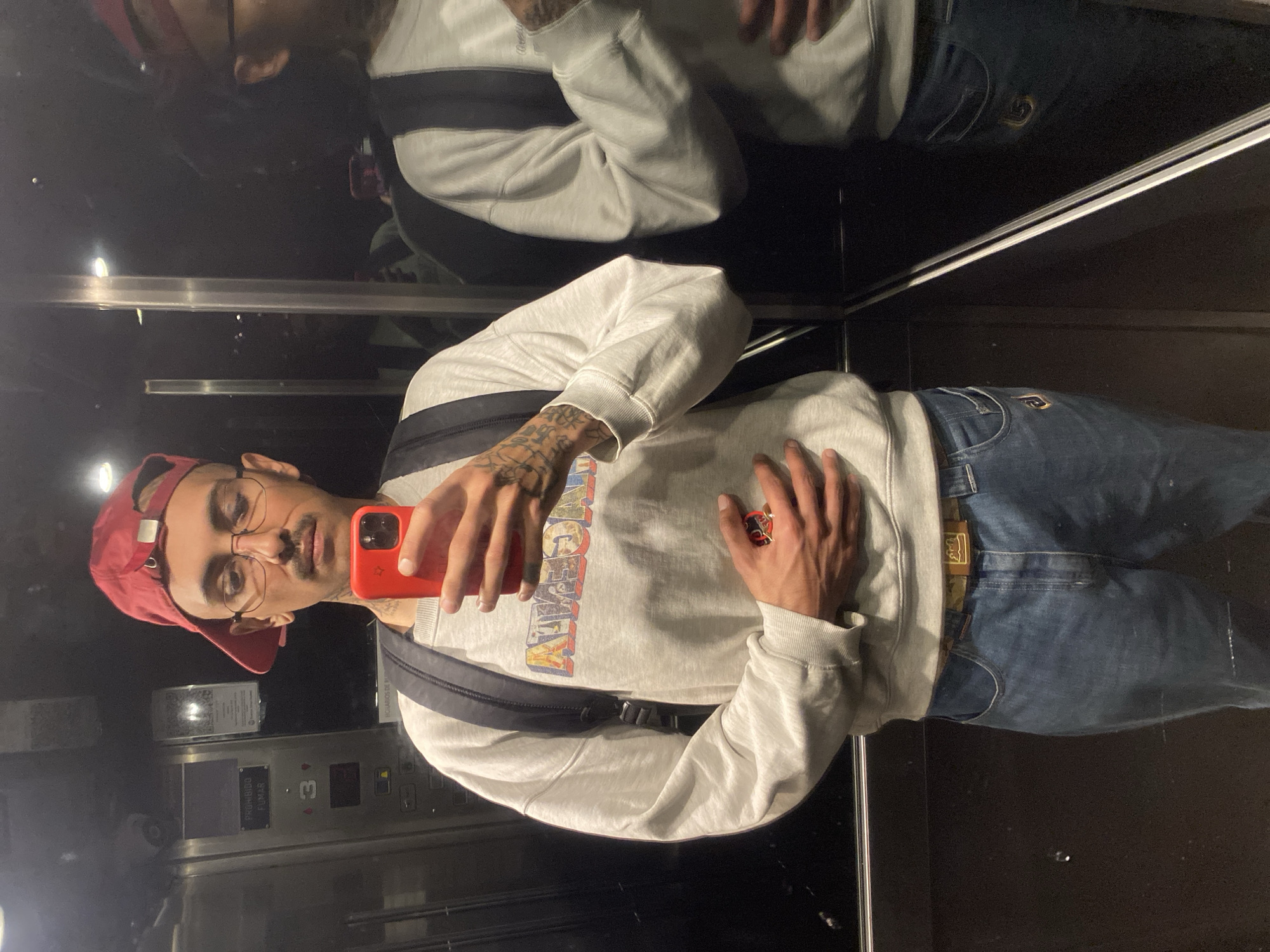 A man is taking a selfie in an elevator. He is wearing a red baseball cap, glasses, a light grey sweatshirt with a graphic design, and blue jeans. He is holding a red smartphone in his left hand. He has tattoos on his left forearm. The elevator has a stainless steel interior and a control panel with buttons numbered 3. The image is likely taken in Buenos Aires, Argentina, given the description of the environment. The man is interacting with his phone to take a photo of himself in the elevator.