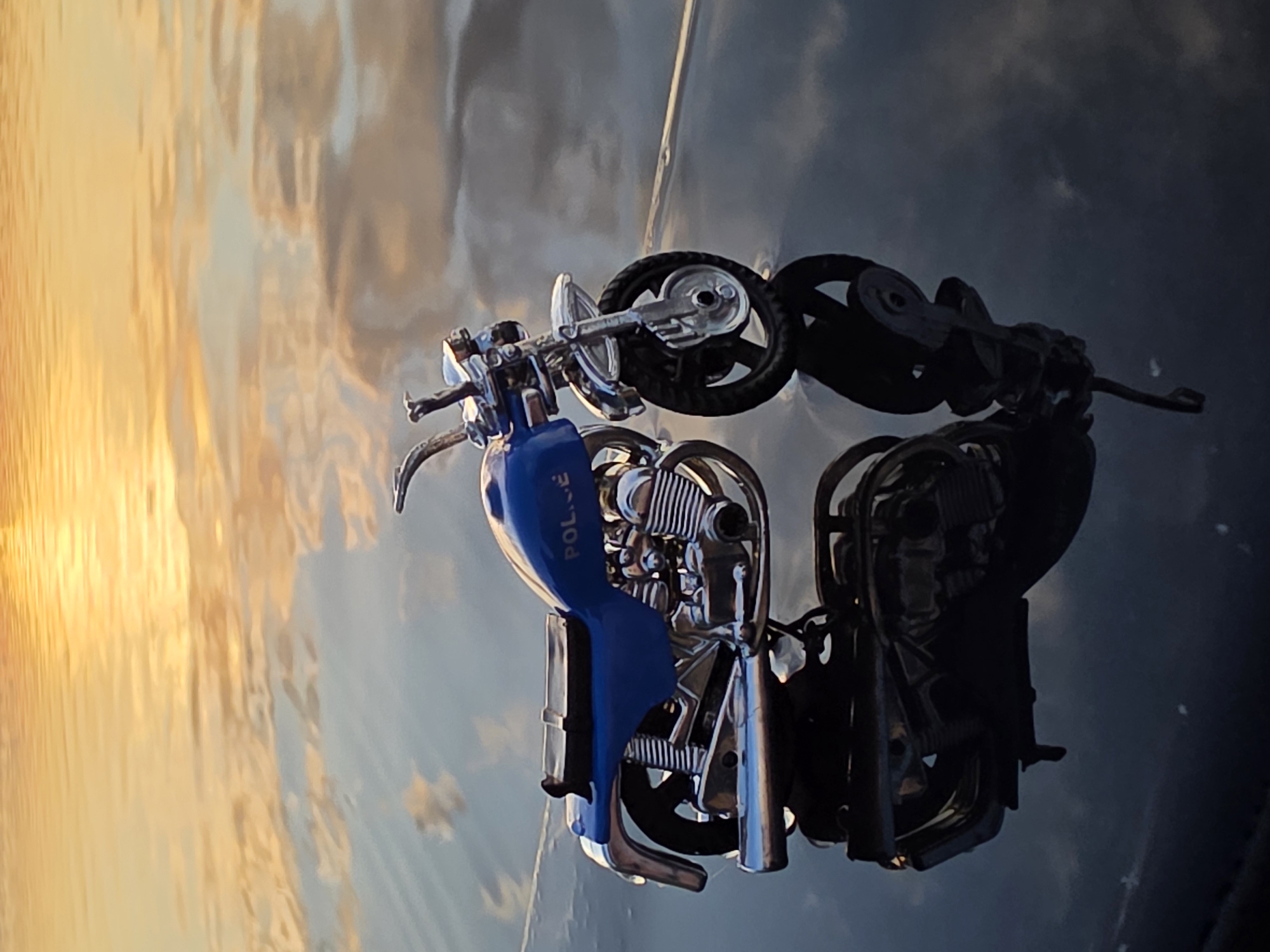 A close-up, eye-level shot shows a toy police motorcycle with a blue gas tank and chrome accents, positioned on a dark, reflective surface, possibly the hood of a car. The motorcycle is seen from the side, with its front wheel on the right. The reflection of the motorcycle is visible on the surface below. In the background, a body of water reflects the golden and orange hues of a sunset or sunrise, with subtle hints of clouds. The scene is located in Chaikovsky, Russia.