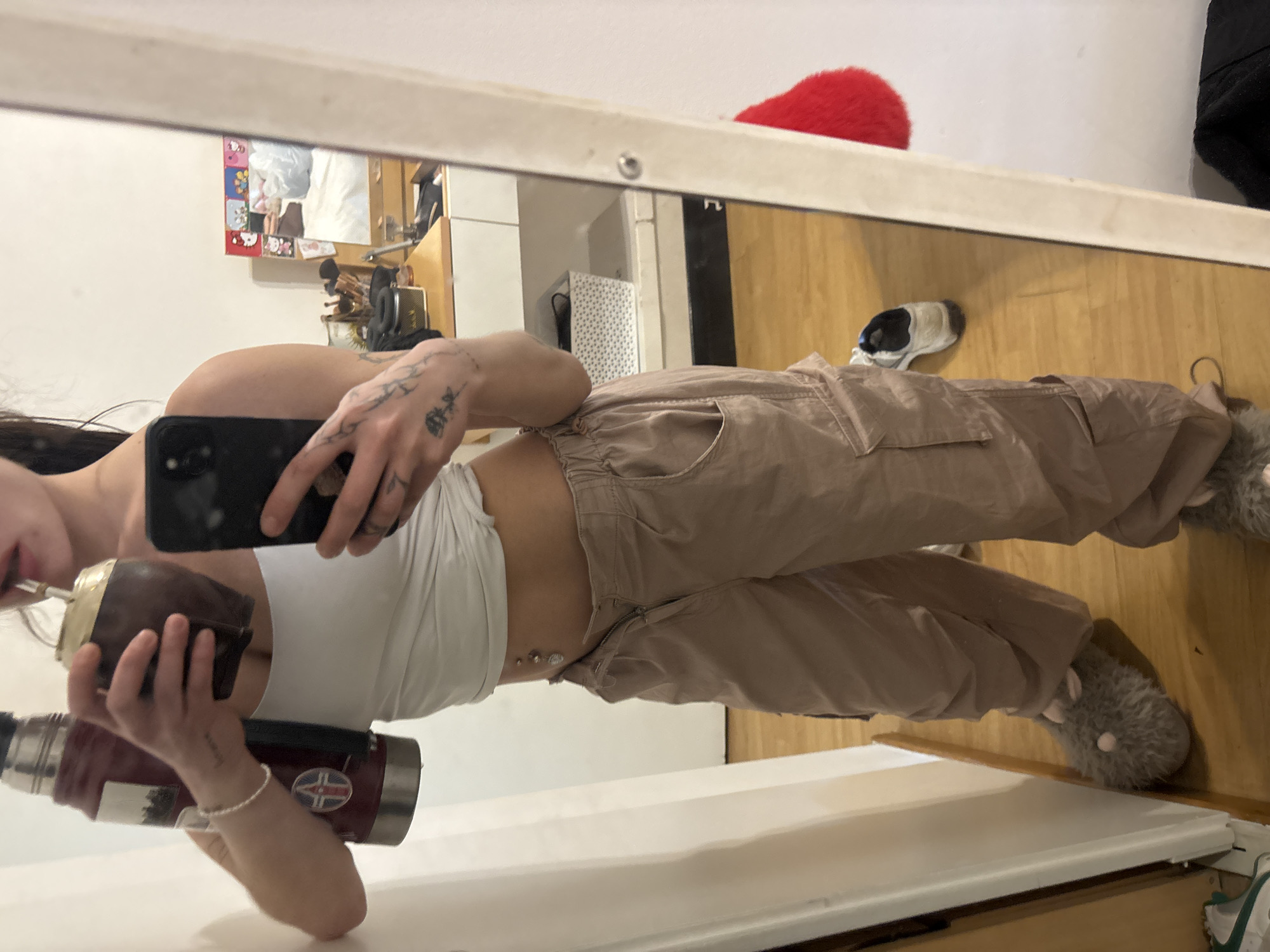 A mirror selfie captures a person reflected in a full-length mirror within an indoor setting. The image is rotated, showing the floor on the right side and a white wall on the left. The subject, a person with long dark hair, is wearing a white crop top, light brown cargo pants, and fluffy grey slippers with white details. They have visible tattoos on their hands and forearms, as well as a navel piercing.
The person holds a black smartphone in one hand, presumably taking the photograph. In the other hand, they hold a dark brown mate gourd with a metal bombilla (straw) and a red thermos bottle, indicating preparation or consumption of mate, a prominent cultural tradition in Buenos Aires, Argentina. The thermos bears a circular logo featuring a white cross on a red background, possibly with blue accents.
The background, visible in the mirror's reflection, reveals a domestic space with a light-colored wooden floor. On the wall, several small pictures, including Hello Kitty designs, are arranged. Other reflected items include a white and black patterned box, a wooden furniture piece, a red fuzzy object (possibly a pillow), and a black and white sneaker. The scene depicts a casual, everyday moment in a home environment. No specific time of day or weather is discernible.