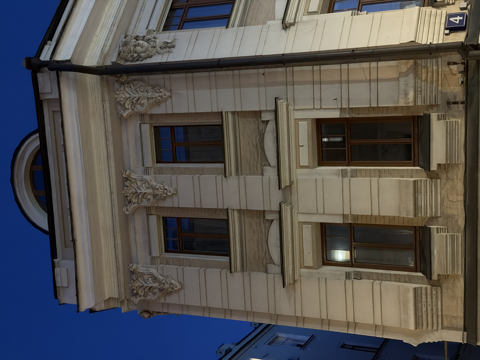 The image shows a building facade in Moscow, Russia. The building has a light-colored facade with ornate decorations around the windows and near the roofline, including sculpted faces and floral patterns. There are several windows with dark frames visible. A drainpipe runs vertically down the building's edge. A blue sign with the number "4" is visible on the upper right side of the building. The sky is a deep blue, suggesting it is dusk or evening.