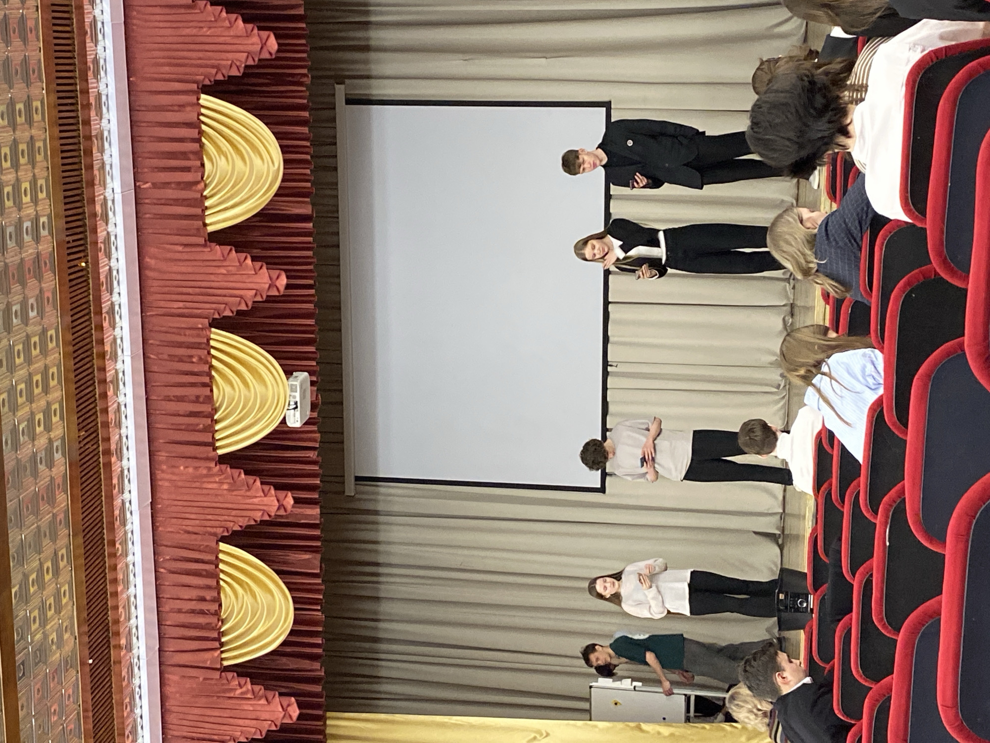 The image depicts an auditorium setting in Votkinsk, Russia. A stage is visible with a large projection screen in the background, flanked by draped beige curtains. Along the left side of the image is a section of the auditorium's decor, featuring a multi-layered design in reds and golds.
Several people are on stage. Two individuals, a male and female, are positioned towards the top of the frame, dressed in formal attire. Further down the stage are a few other individuals in more casual clothing.
In the foreground, to the right, are rows of auditorium seats upholstered in red and black. Occupying these seats are audience members of various ages and attires. A male individual is seated at the very bottom of the image, near the stage, in a dark jacket or suit.