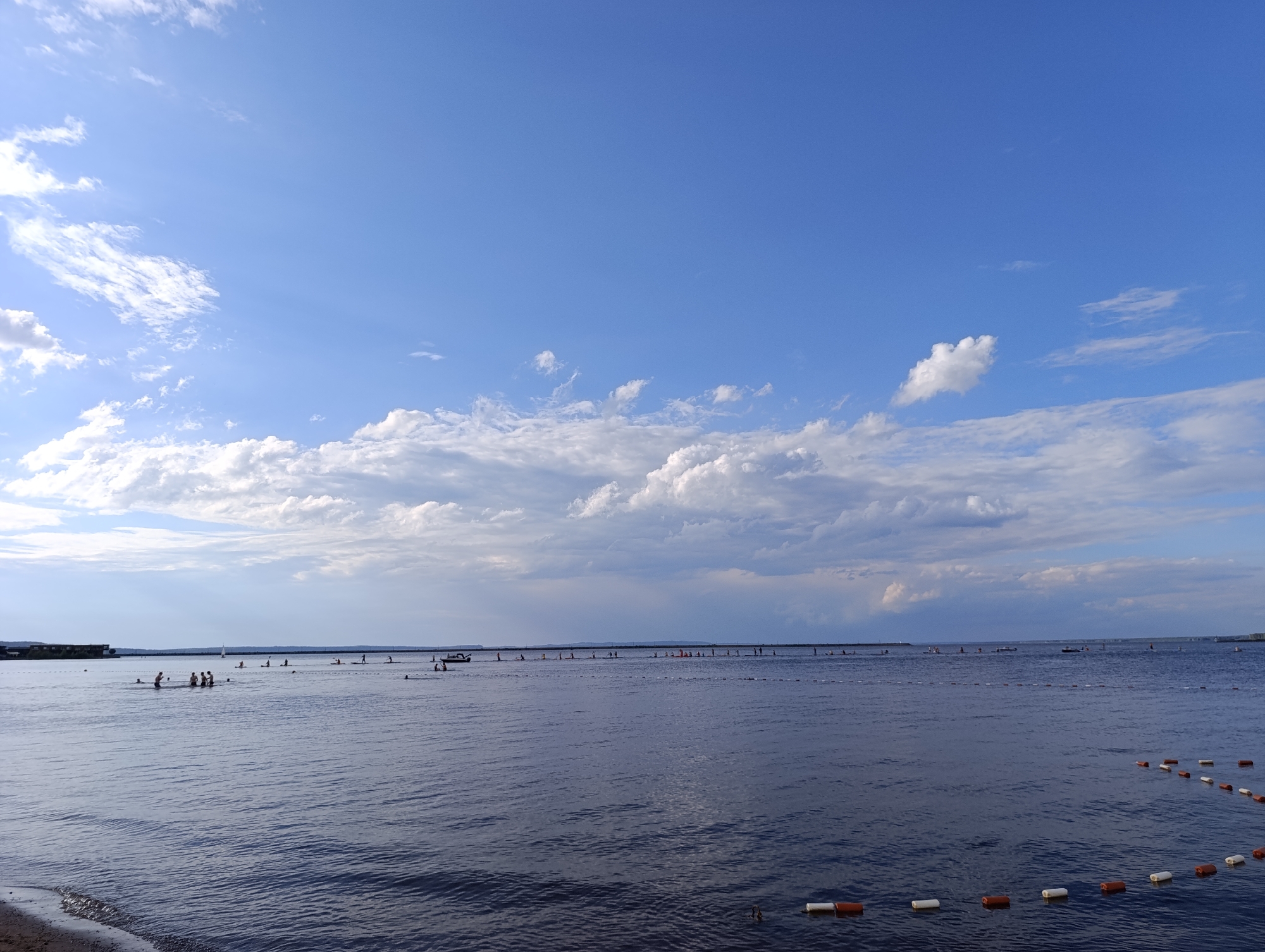 This is a wide shot of a calm body of water under a partly cloudy blue sky. In the foreground, the water is dark and reflects the sky, with small waves lapping against a sandy shore on the left. In the middle ground, there are numerous people scattered across the water, some appearing to be swimming or wading, while others are standing on what look like buoys or small rafts. Further in the distance, a line of buoys stretches across the water, with more people visible along it, some appearing to be engaged in water sports like paddleboarding or kayaking. A small boat is also visible in the distance. The background shows a long, flat shoreline with some buildings or structures. The overall scene suggests a recreational day at the beach or a lake during daylight hours, likely in the late afternoon given the lighting. The weather appears pleasant and suitable for outdoor water activities.