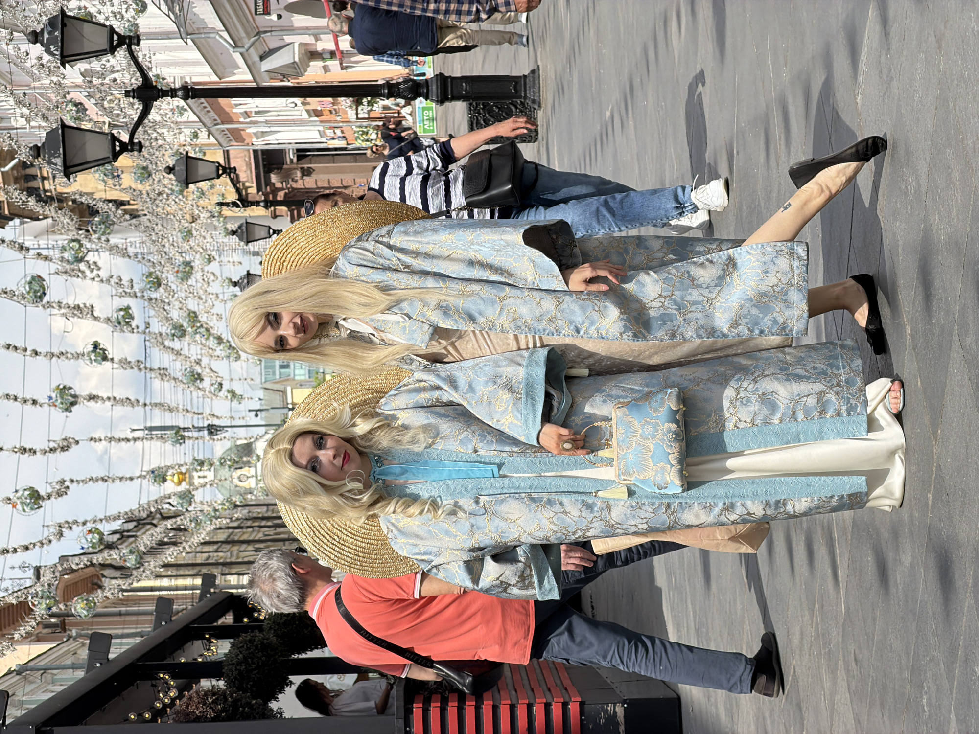 The image captures a vibrant daytime street scene in Moscow, Russia, under bright, clear skies. In the foreground, two women with long blonde hair are the central subjects. Both are dressed in striking, elaborate long coats of light blue fabric adorned with intricate gold patterns. They each wear a large, wide-brimmed straw hat and black flat shoes. The woman on the right has a small tattoo visible on her ankle, and one of the women carries a small, decorative handbag that matches her coat. Their distinctive attire suggests they may be performers or participants in a special event.
The background reveals a bustling urban pedestrian area. The street is decorated with ornate black street lamps featuring multiple light fixtures, and overhead, numerous sparkling, spherical ornaments are strung across the street, creating a festive atmosphere. The ground is a uniform paved surface, reflecting the daylight.
Other individuals are visible throughout the scene. On the left, closer to the foreground, a man in a vivid orange polo shirt and dark trousers walks with a black messenger bag; the brand name "RITTER" is clearly discernible on its strap. Further back, another person in a striped shirt and blue jeans is seen. The street is lined with various buildings, displaying different architectural styles, hinting at an established city environment. A blurry sign in the far background might contain the Russian text "ТАСКА". The overall impression is that of a cheerful and active public space.