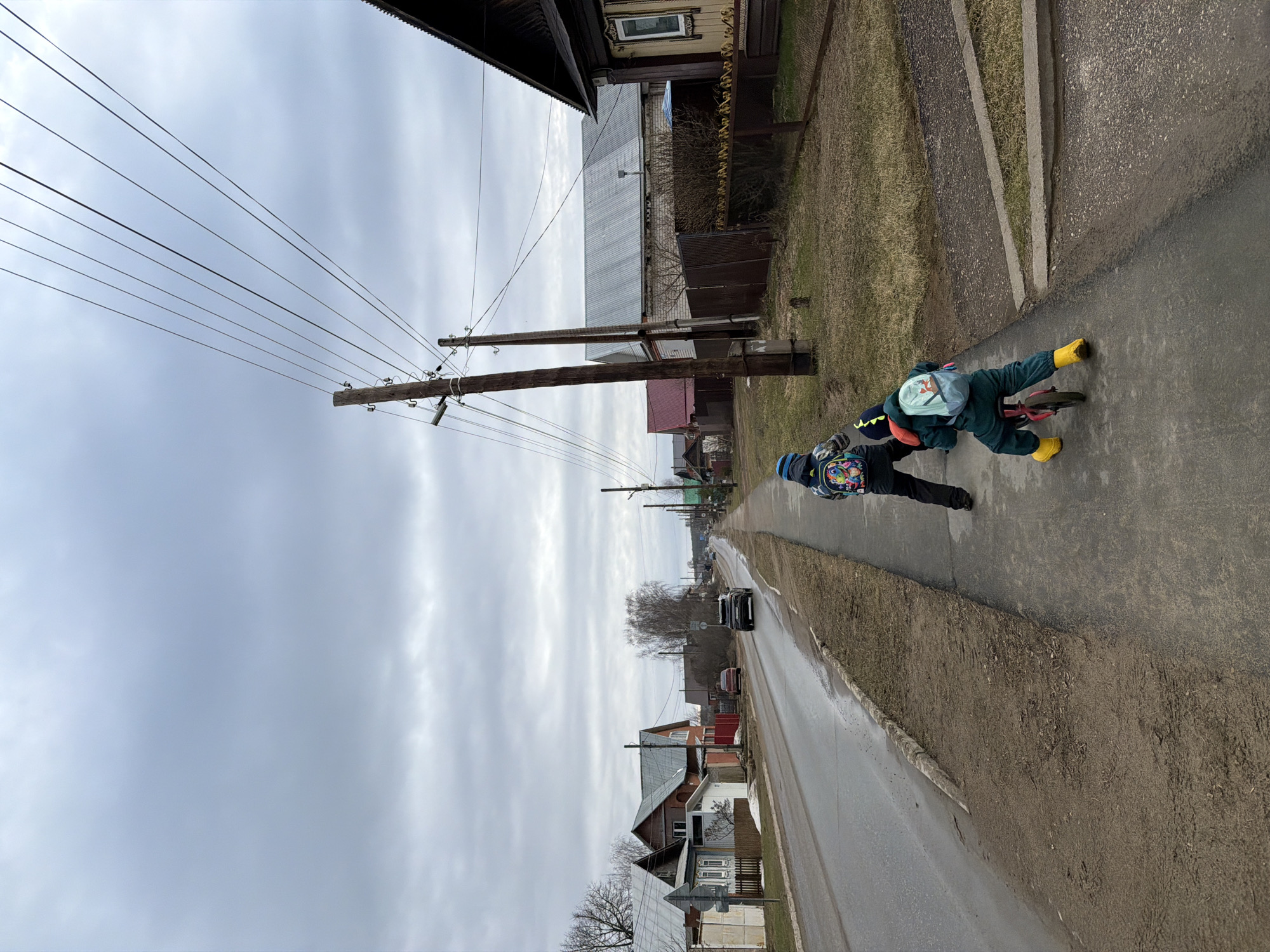 A street scene in Chaikovsky, Russia depicts two individuals, likely children, on a sidewalk. One is riding a bicycle with yellow boots, and the other is walking. The street is wet, and there is a car in the background. Power lines run along the street, and traditional Russian houses line the road. The sky is overcast.