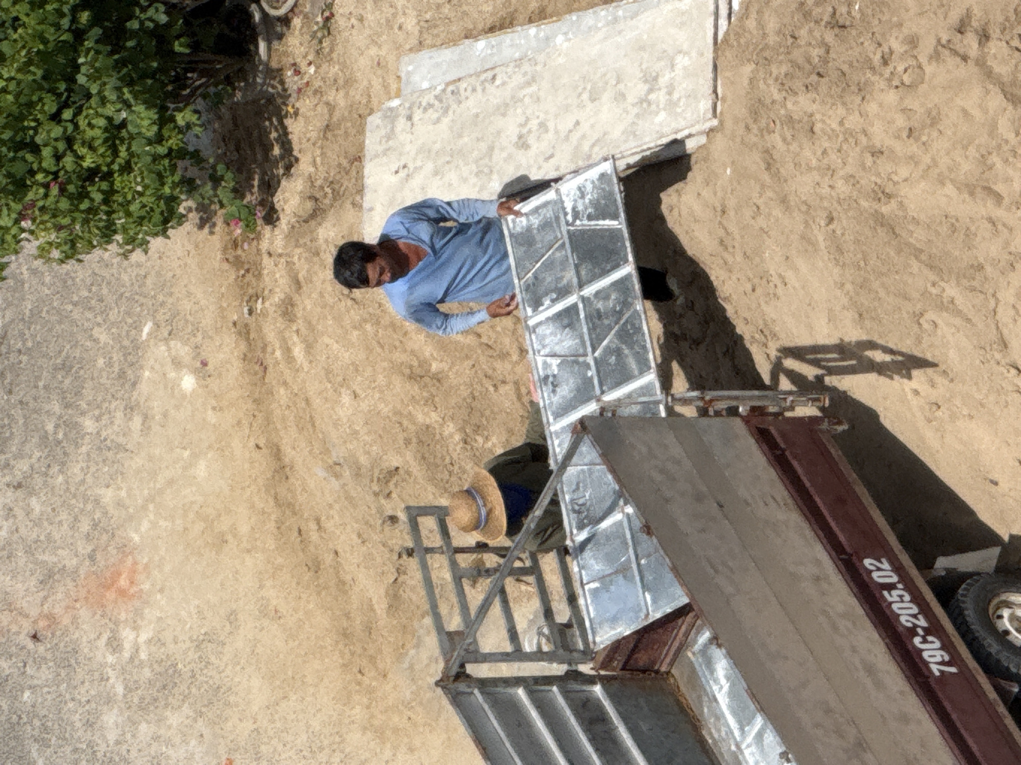 The image depicts an outdoor scene in Bac Nha Trang Ward, Vietnam, likely during the daytime. Two individuals are visible, engaged in some form of construction or loading activity. One person, wearing a light blue long-sleeved shirt and dark pants, is standing and holding a large, flat, metallic object that appears to be a window frame or a similar construction panel. This individual has dark hair and is looking downwards.
The second person is partially visible, wearing a straw hat with a blue band and a dark-colored garment. They are positioned near a metallic structure that resembles a ramp or part of a trailer. This metallic structure is connected to a larger, dark-colored trailer with wooden planks visible on its surface. A tire from the trailer is also visible.
The ground is a mix of sand and concrete, with some vegetation in the upper left corner of the image, suggesting an outdoor work site. Shadows are cast on the sand, indicating it is likely sunny. The overall impression is of manual labor taking place outdoors.
A visible piece of text on the side of the trailer reads "79C-205.02" in white lettering. The overall scene suggests a busy, functional environment rather than a leisure setting.
