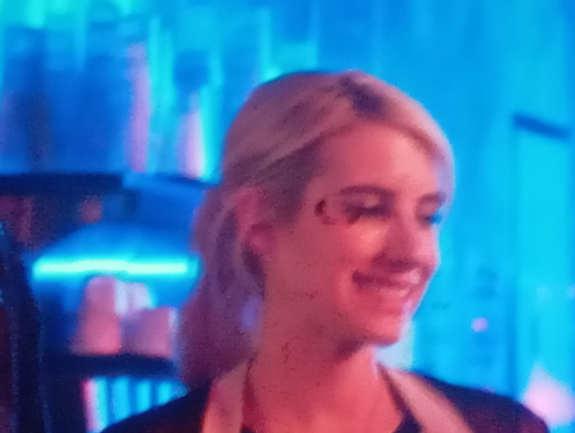 The image is a close-up, low-resolution, and slightly grainy photograph of a young woman with blonde hair pulled back in a ponytail. She is smiling and looking slightly to her left. A subtle, crescent-moon-shaped marking is visible on her left cheekbone. She is wearing a dark garment, possibly a shirt or vest, over another lighter-colored garment.
The background is blurred and consists of bright, vertical blue and pink neon lights. In the lower left portion of the image, blurred objects that appear to be pink cups or containers are visible on shelves, illuminated by the neon lights. The overall environment suggests a dimly lit, possibly commercial space with vibrant lighting. The image does not provide any discernible information about the city of Sofiivska Borshchahivka, Ukraine.