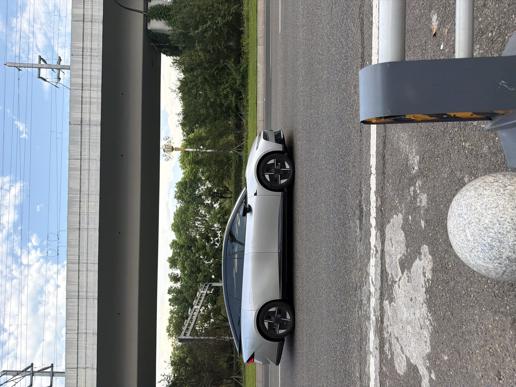 An eye-level, medium shot captures a silver sedan with a black roof and distinctive black and silver wheels driving on a paved road. The car is positioned in the right half of the frame, facing left. The road surface is asphalt, with a white lane marker visible. In the foreground, to the right, a grey and yellow traffic barrier is partially visible, along with a grey granite sphere to the left. Behind the car, a line of dense green trees forms a backdrop. Above the trees, a concrete overpass stretches horizontally across the upper portion of the image. Overhead power lines are suspended above the overpass. The sky is visible above the power lines, with scattered white clouds against a pale blue expanse. The scene appears to be in a semi-urban or suburban environment, possibly along a highway or major road. The specific location is Jiangjin District, Chongqing, China.