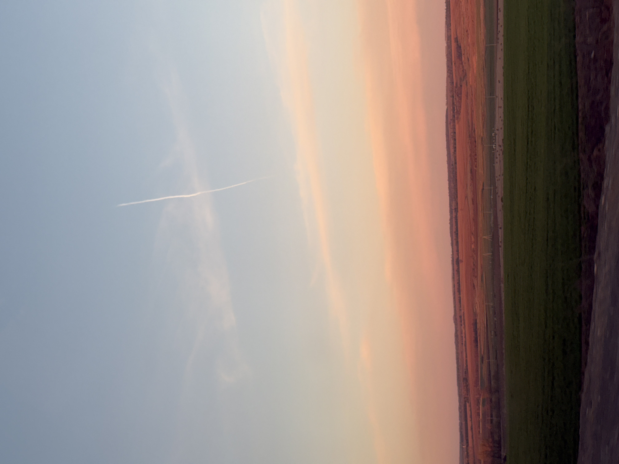 The image depicts a view of the landscape in Камаево, Russia, featuring a sky with hues of blue, white, and peach, possibly during sunrise or sunset. A faint contrail cuts across the blue portion of the sky. Below the sky, the landscape shows fields of green and brown, possibly farmland or natural terrain. The image appears to be taken from a high vantage point, slightly rotated. No people, specific objects, or interactions are visible.