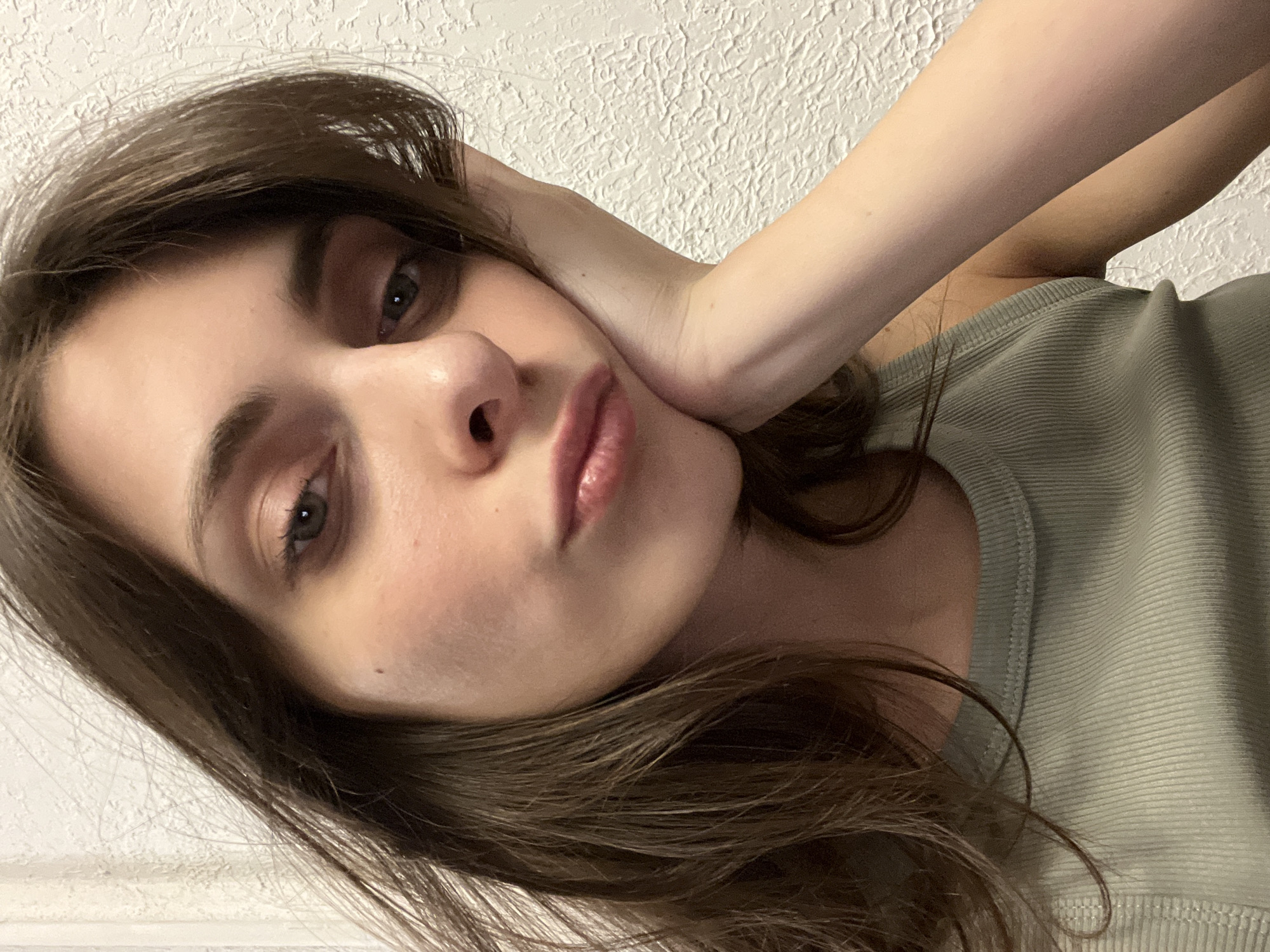 A young woman with brown hair and green eyes is posing for a selfie. Her left hand is pressed against her face. She is wearing a green ribbed top. The background is a textured white wall. The image does not provide any information to suggest it was taken in Krasnoyarsk, Russia.