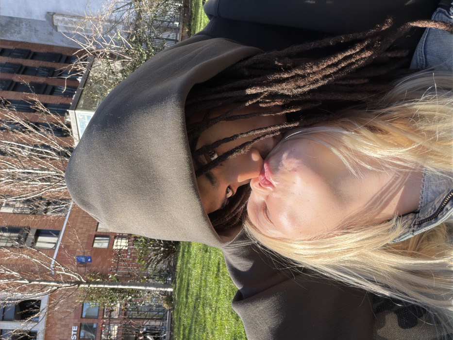 The image depicts a close-up, chest-up outdoor portrait of two individuals kissing. On the right, a person with dark dreadlocks or braids wears a dark olive-green or dark grey hooded sweatshirt, with the hood pulled up, partially obscuring their face. Their right eye is visible, looking directly towards the camera. On the left, a blonde-haired individual with an upward-tilted head and closed eyes is kissing the hooded person, their lips pursed. This individual wears a light-colored, possibly denim, jacket. The action is an intimate interaction between the two subjects.
The scene is set in a public park or green space within an urban area of Paris, France. The background features bright green grass and several bare-branched trees, suggesting a cooler season. Beyond the green space, urban architecture is visible. A prominent multi-story building with a reddish-brown brick facade and dark-framed windows stands behind a black wrought-iron fence. A white commercial sign bearing "STE" (part of "Lacoste") and its distinctive green crocodile logo is visible on this building. A blue rectangular public information plaque, characteristic of Parisian street signage, is also present on a pole or fence. Further in the background, a tall, light-colored building with vertical textures is visible. The entire scene is illuminated by strong natural sunlight.