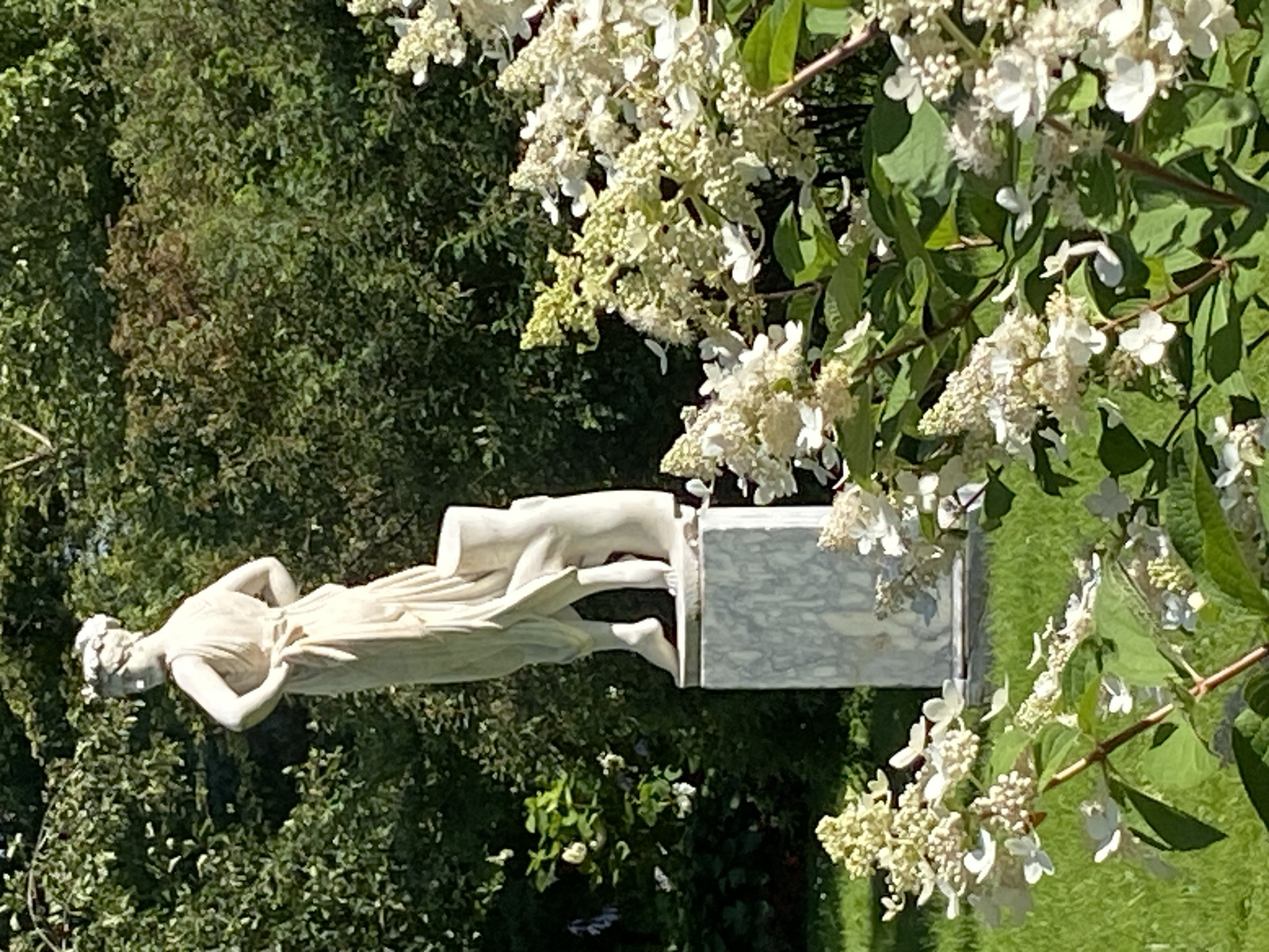 A full-shot, eye-level outdoor photo captures a classical marble statue of a woman in a garden. The statue depicts a woman in a draped gown with her hands on her hips, standing next to a tree stump. She is on a rectangular marble pedestal. In the foreground, white flowers with green leaves are in bloom. Behind the statue, a dense green forest serves as the backdrop. The lighting suggests it is daytime, with sunlight illuminating the scene and casting shadows. The image appears to be taken in Korpikiulya, Russia.