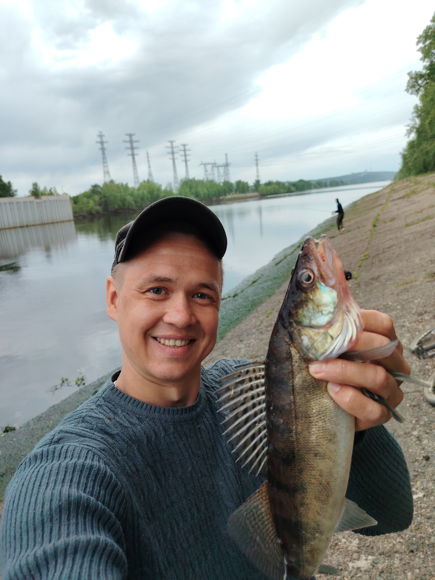 A man holds a fish while standing next to a canal in the city of Chaikovsky, Russia. The man is fair-skinned, with short hair and a cap, and he is smiling. He is wearing a blue-gray sweater. The fish is silvery-brown with a distinct dorsal fin. In the background is a canal with concrete sides, water, green trees on the opposite bank, and power lines in the distance. Another person is fishing in the background. The sky is overcast.
