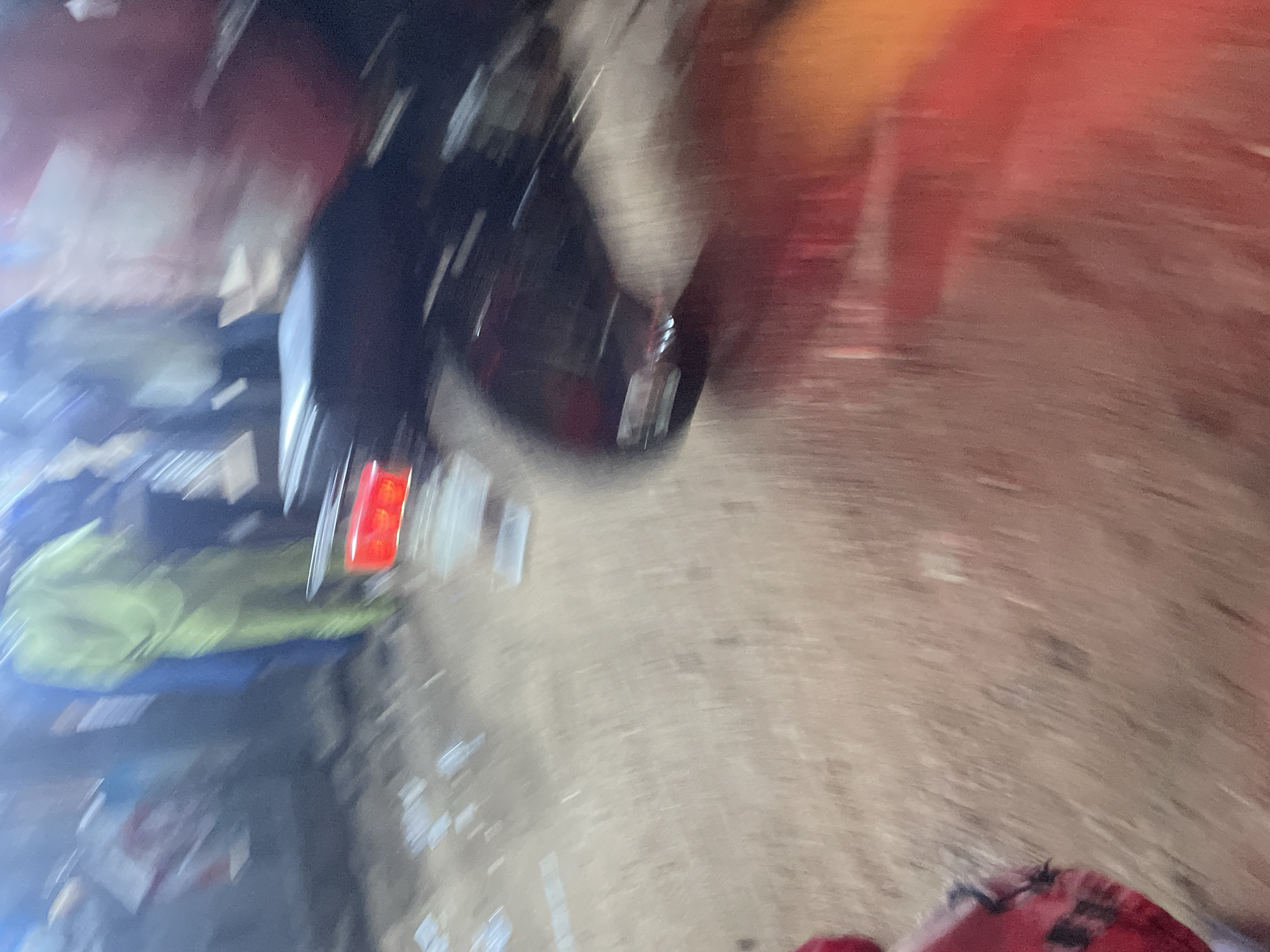The image captures a scene that appears to be on a street in Lokoja, Nigeria, likely at night or in low light conditions, given the motion blur and artificial lighting. A motorcycle, possibly a commercial one, is central to the composition, its red taillight illuminated. The motion blur suggests movement of the motorcycle or the camera. In the background, several individuals are visible, some wearing bright, possibly protective clothing, like a person in a neon green garment, and others in darker attire. The ground appears to be a mixture of dirt and possibly debris. The overall impression is of a busy, perhaps dynamic, urban environment.