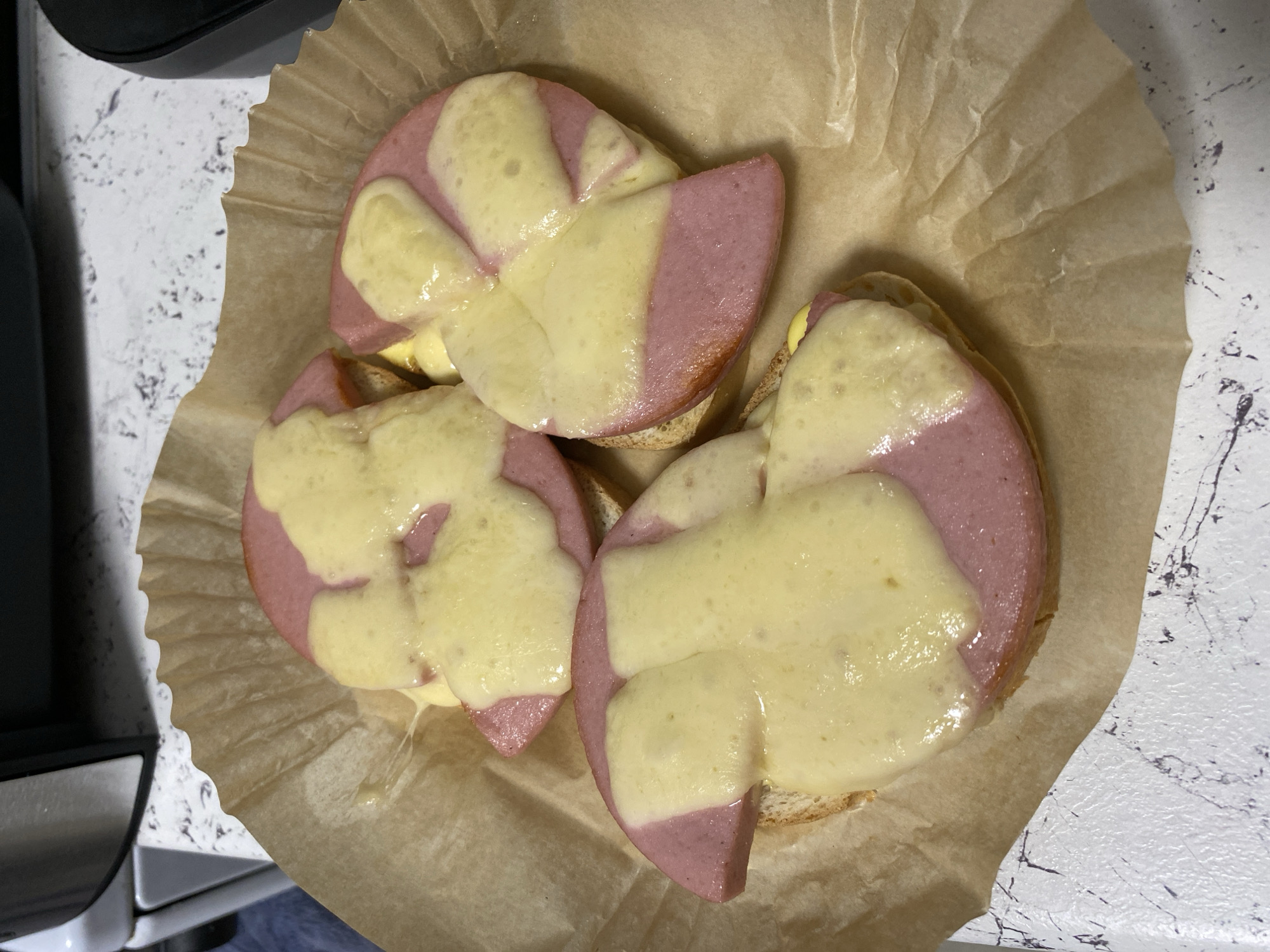 The image shows three open-faced sandwiches on a crumpled piece of brown parchment paper. Each sandwich consists of a slice of round bread topped with a thick slice of bologna. Melted cheese covers a large portion of each bologna slice. The sandwiches are arranged close together on the paper, which is placed on a countertop with a speckled gray and white pattern. A black object is partially visible in the upper left corner of the image. The image provides no location information related to the city of Noviy, Russia.