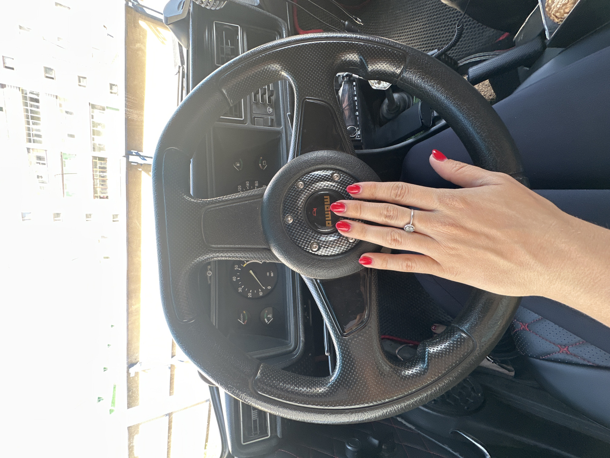 The image shows a close-up view of a person's hand resting on the steering wheel of a car. The hand has bright red nail polish and a diamond ring on the left ring finger. The steering wheel is black and has a textured grip. The car's dashboard is visible, with gauges and a stereo system. The background shows a bright, sunlit view through the windshield, with buildings and some trees. The scene appears to be in a city, with the light suggesting it is daytime. Based on the metadata associated with the image, the location is Novoaltaysk, Russia.