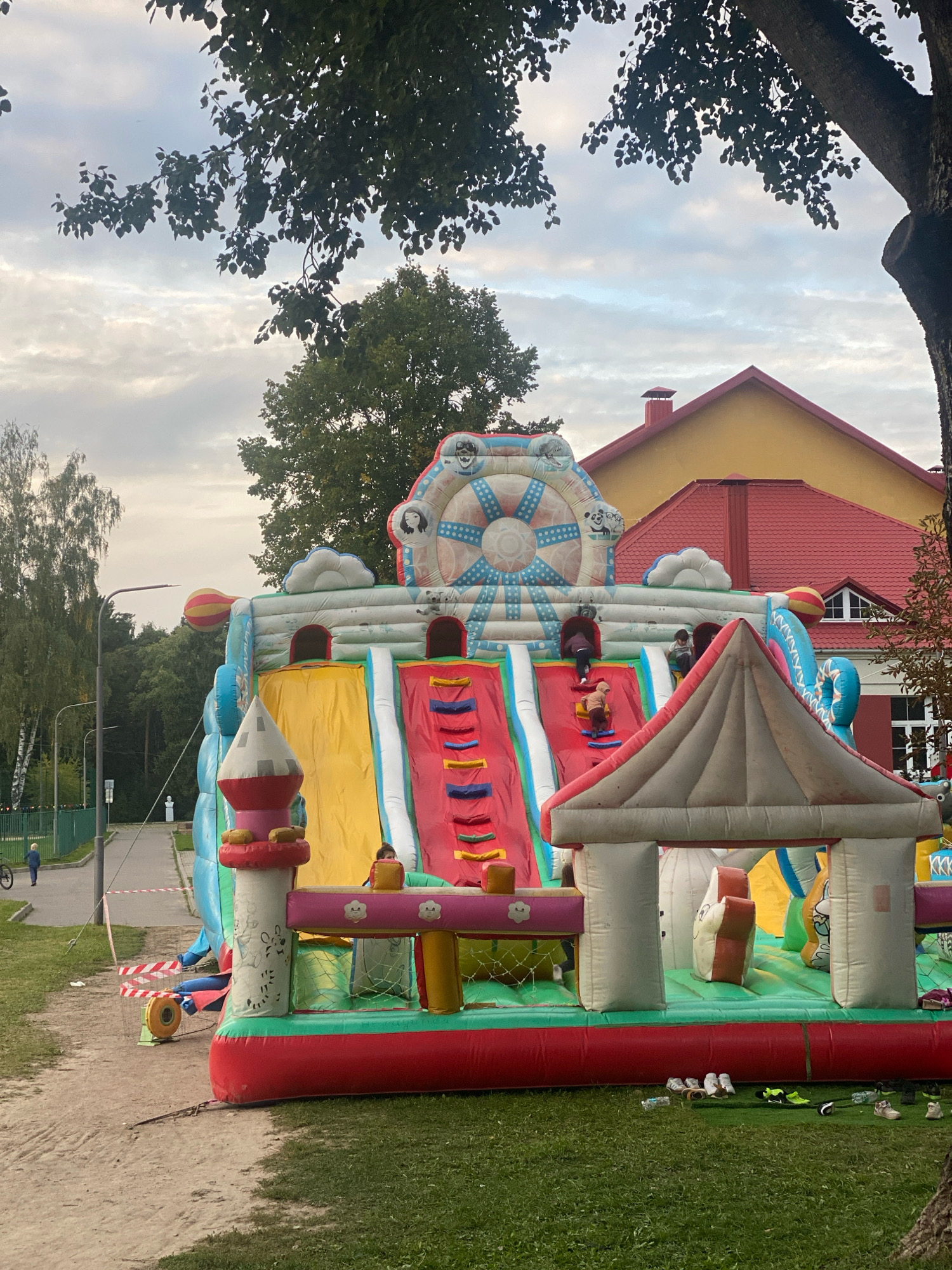 An inflatable playground in Machulishchy, Belarus, features a large, colorful structure with slides and climbing areas. The playground is situated on a grassy area adjacent to a sandy path. Several children are visible on the inflatable, either climbing or descending the slides. In the background, a yellow building with a red roof stands, and trees surround the area. A street with a lamppost and a green fence is to the left, with a person on a bicycle and another walking further down the path. Red and white caution tape is visible near the playground, and various shoes are scattered on the grass in front of it. The sky is overcast with clouds.