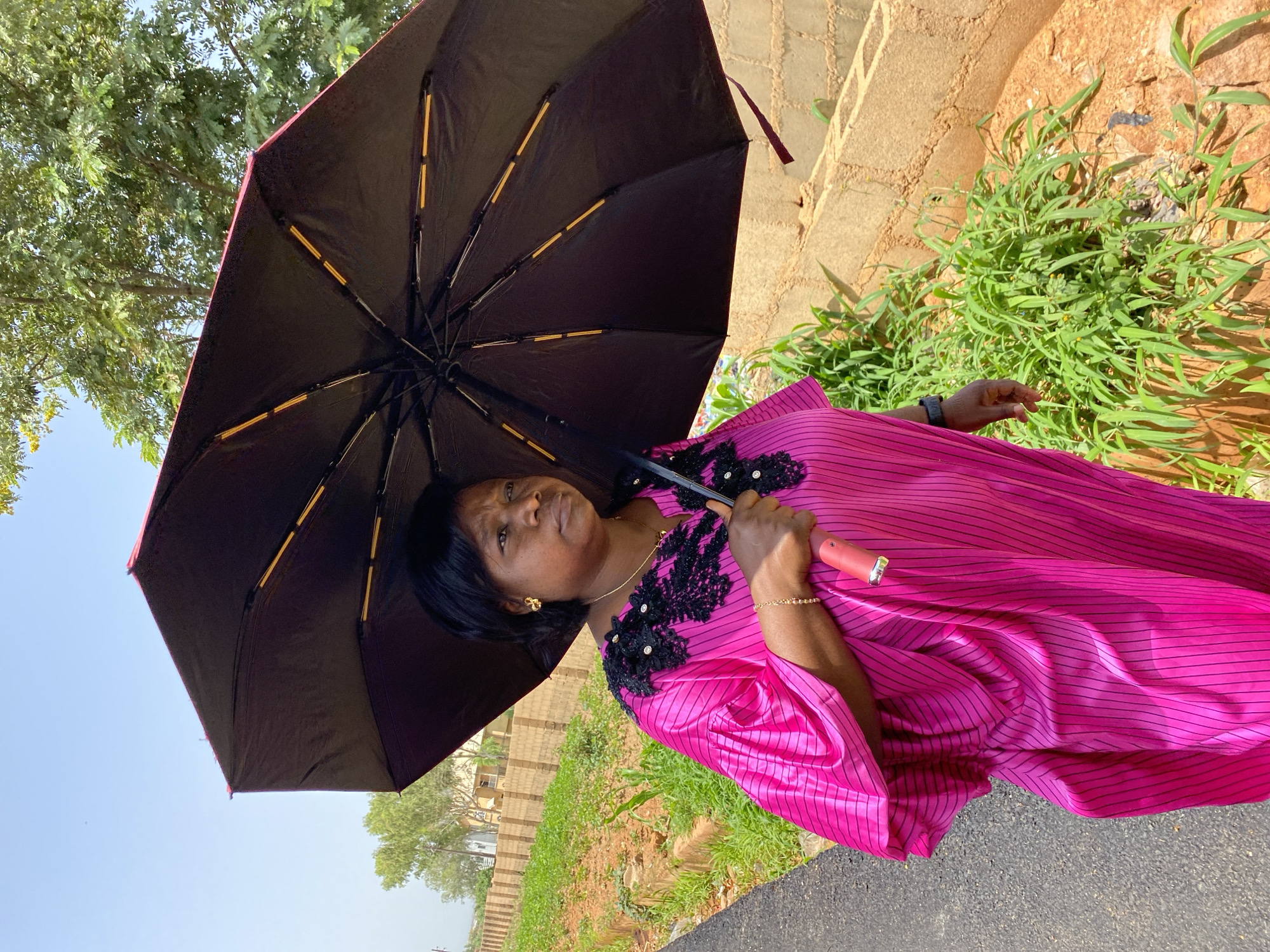 A woman stands outdoors holding a partially opened black umbrella with a red trim. She is wearing a vibrant pink dress with black pinstripes and black lace detailing around the neckline. She has dark hair and is wearing gold earrings and a delicate gold bracelet. Her expression appears contemplative. The scene is set in a sunny environment, suggested by the bright sky and the woman holding an umbrella. Green vegetation and a textured, sandy-colored pathway or wall are visible in the background, along with hints of buildings. The ground surface in the foreground appears to be asphalt. The overall impression is of a person seeking shade on a bright day, likely in a location like Bukuru, Nigeria, given the context. No visible text is present in the image.