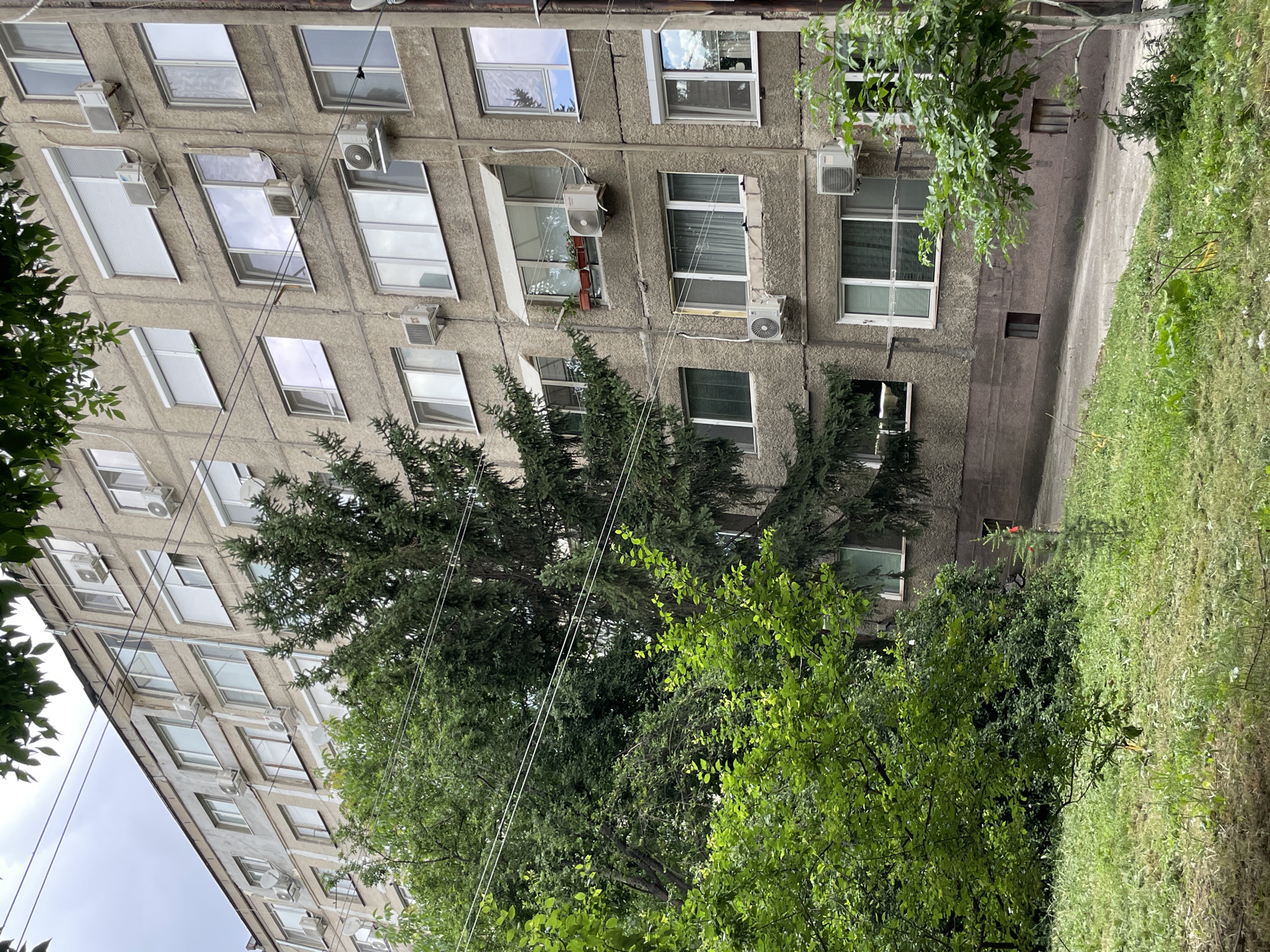 The image shows a close-up of a multi-story apartment building with a concrete facade. The building has numerous windows, many of which are fitted with air conditioning units. There are also several roller shutters visible on some windows. Lush green trees and foliage partially obscure the building, with some branches and leaves hanging into the frame from the top and sides. Power lines are also visible crisscrossing the upper portion of the image. The ground in front of the building is covered in grass and a paved walkway. The overall impression is of a residential area within a city, with a mix of urban infrastructure and natural elements.