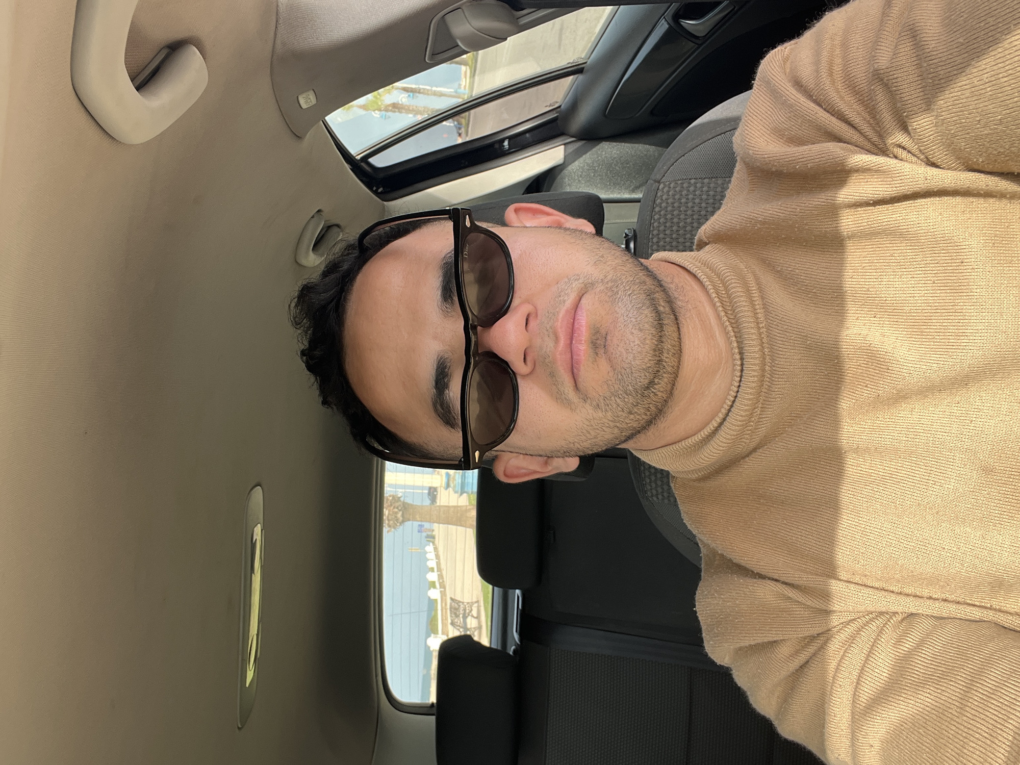 The image displays a man inside a car, photographed in a rotated orientation, appearing 90 degrees counter-clockwise from upright. The main subject is a man, shown from the chest up, with dark hair and light stubble. He wears dark-tinted sunglasses, with the brand "Dior" clearly visible on the temple, and a light brown or beige ribbed knit sweater. His expression is neutral and relaxed.
The setting is the interior of a car. Details of the car include a light-colored headliner with a grab handle and an "AIRBAG" warning label. Dark fabric car seats are visible behind and beside the man. An open sunroof is present, allowing ample natural light to enter and revealing a glimpse of the sky.
Through the car's windows, a bright, sunny outdoor scene is visible. This includes what appears to be a body of water (suggesting the sea or a large bay/river), distant structures, and some trees, indicative of a coastal or waterside location. The strong sunlight indicates clear, daytime weather, casting distinct shadows within the car. The activity is the man posing for a photo, likely a selfie, in a casual moment. The location context is Martil, Morocco.