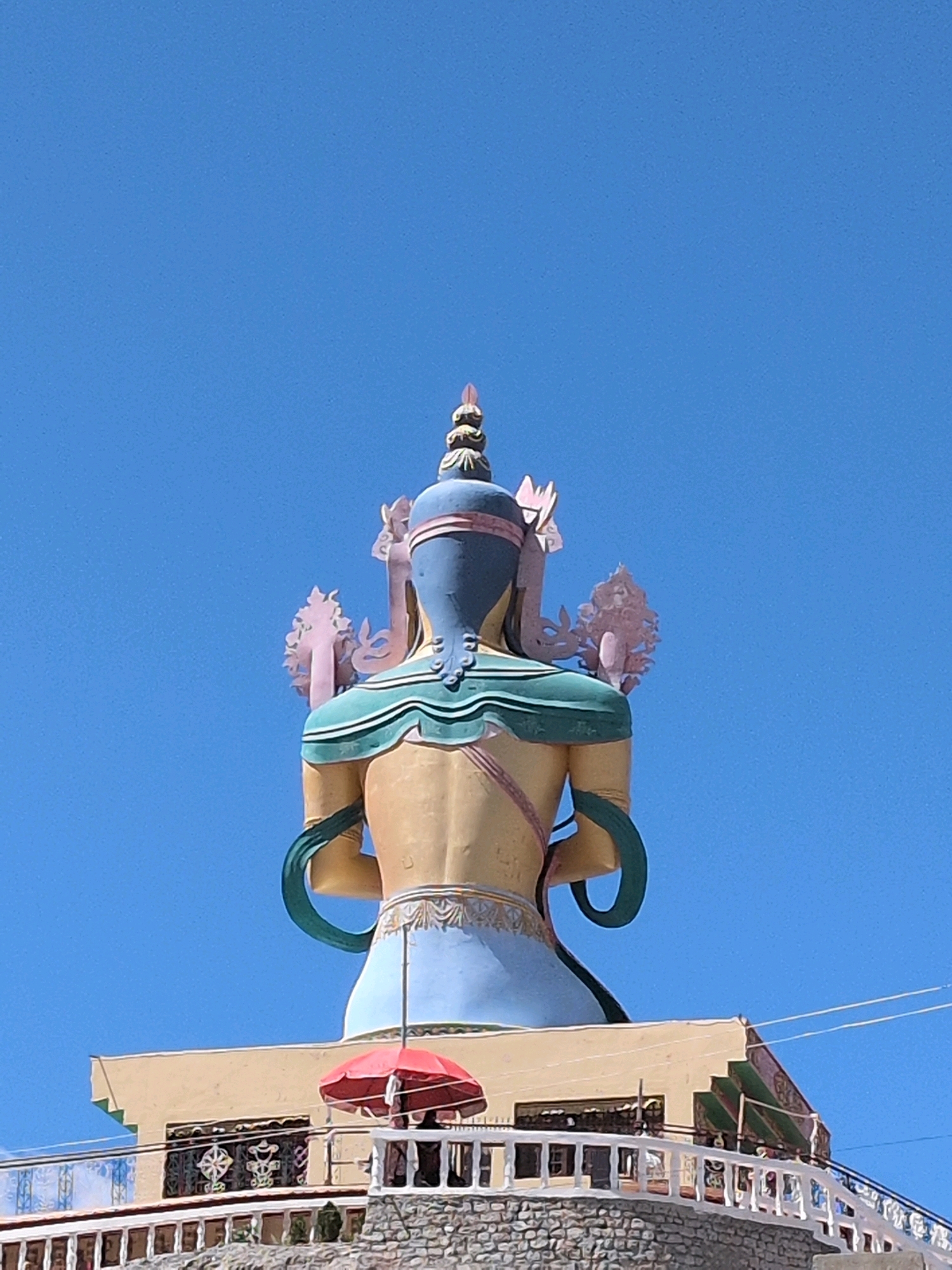 This is a vertical, outdoor, eye-level shot of a large, colorful statue of a deity, seen from behind, against a clear blue sky. The statue is predominantly yellow and blue, with accents of pink and green. It is adorned with elaborate headdresses and jewelry. Below the statue, there is a light yellow building with a red umbrella visible. The building is situated on a stone foundation and has a white railing. The location is identified as Diskit, India, likely a Buddhist monastery given the statue's appearance. No people are clearly visible in the frame.