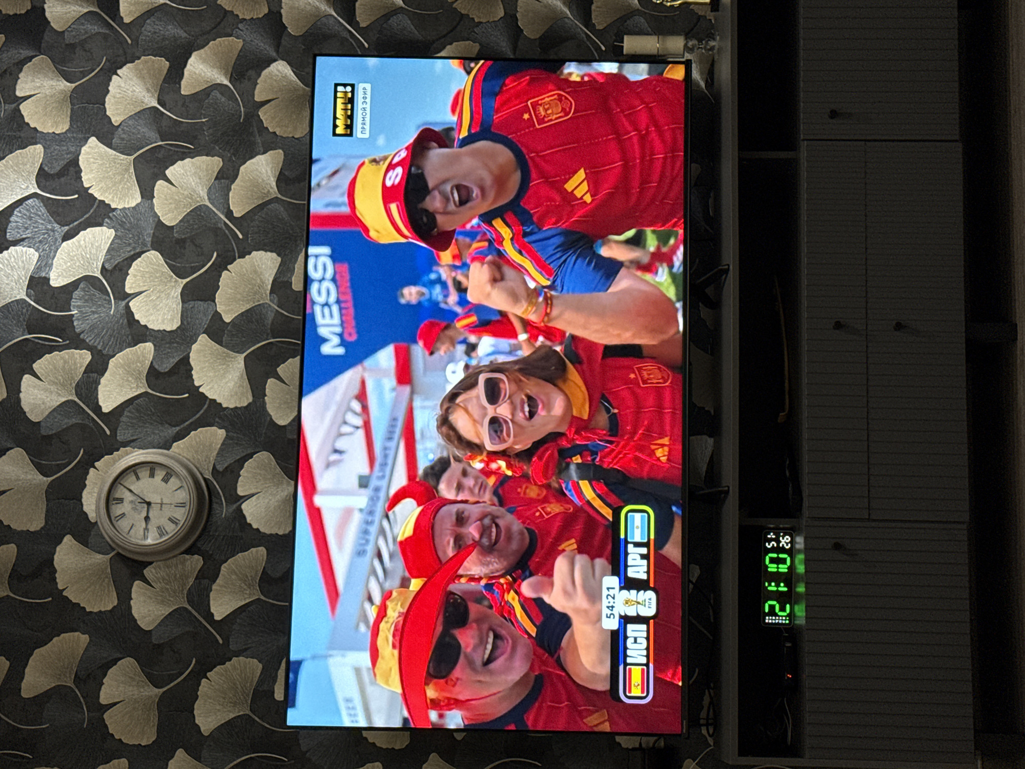 The image shows a television screen displaying a football match between Spain and Argentina, with the score at 54:21. Several fans wearing Spanish national team colors are visible in the foreground, cheering and celebrating. The television is placed on a gray media console, which also has a digital clock displaying the time 21:10 and a temperature of 26 degrees Celsius. In the background, a patterned wallpaper with ginkgo leaf motifs and a clock are visible. The scene appears to be a living room setting, likely in Malmyzh, Russia, given the context of the user's request.