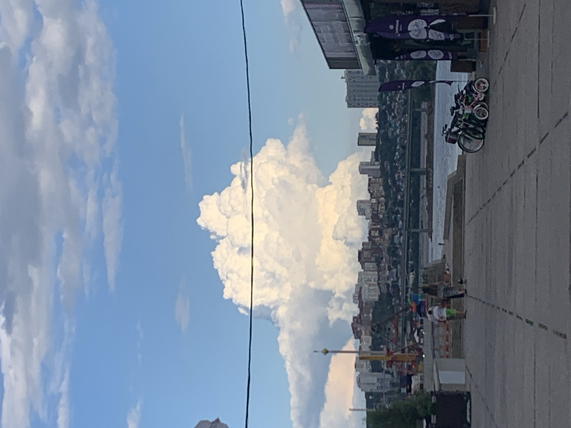 The image depicts a wide paved area in an urban waterfront setting. In the foreground, there are several bicycles parked near a building with purple flags displaying bicycle imagery. Two individuals, a man and a woman, are walking away from the viewer. In the midground, a body of water, likely a river, is visible with a bridge spanning across it. The background features a cityscape with numerous buildings and a large, dramatic cloud formation dominating the sky. The location is identified as the embankment in Ufa, Russia.